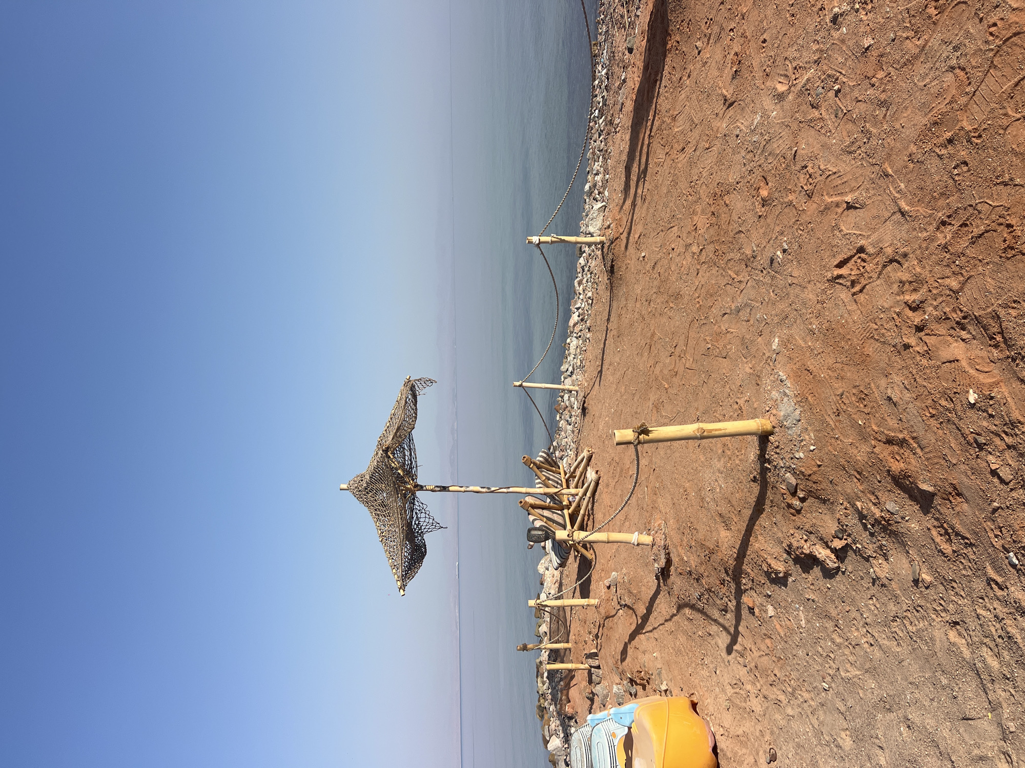 The image depicts a beach scene in Nuweiba, Egypt. A straw umbrella provides shade over two empty lounge chairs made of bamboo and striped cushions. The ground is covered in reddish-brown sand, with scattered rocks and footprints. A rope fence, supported by bamboo poles, separates the sandy area from the rocky shoreline. The calm, blue water of the sea stretches to the horizon, meeting a clear, pale blue sky. On the left side of the frame, a yellow and blue plastic children's toy is partially visible. The overall scene suggests a serene and undeveloped beach environment.