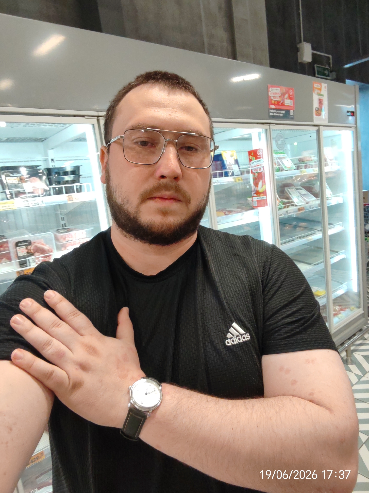 A man with a beard and glasses is standing in front of refrigerated display cases in what appears to be a grocery store. He is wearing a black t-shirt with an Adidas logo. He is holding his left arm with his right hand, which is wearing a watch. The date and time stamp on the image indicates June 19, 2026, at 17:37. The environment suggests a retail setting, likely a supermarket, with products visible in the display cases.