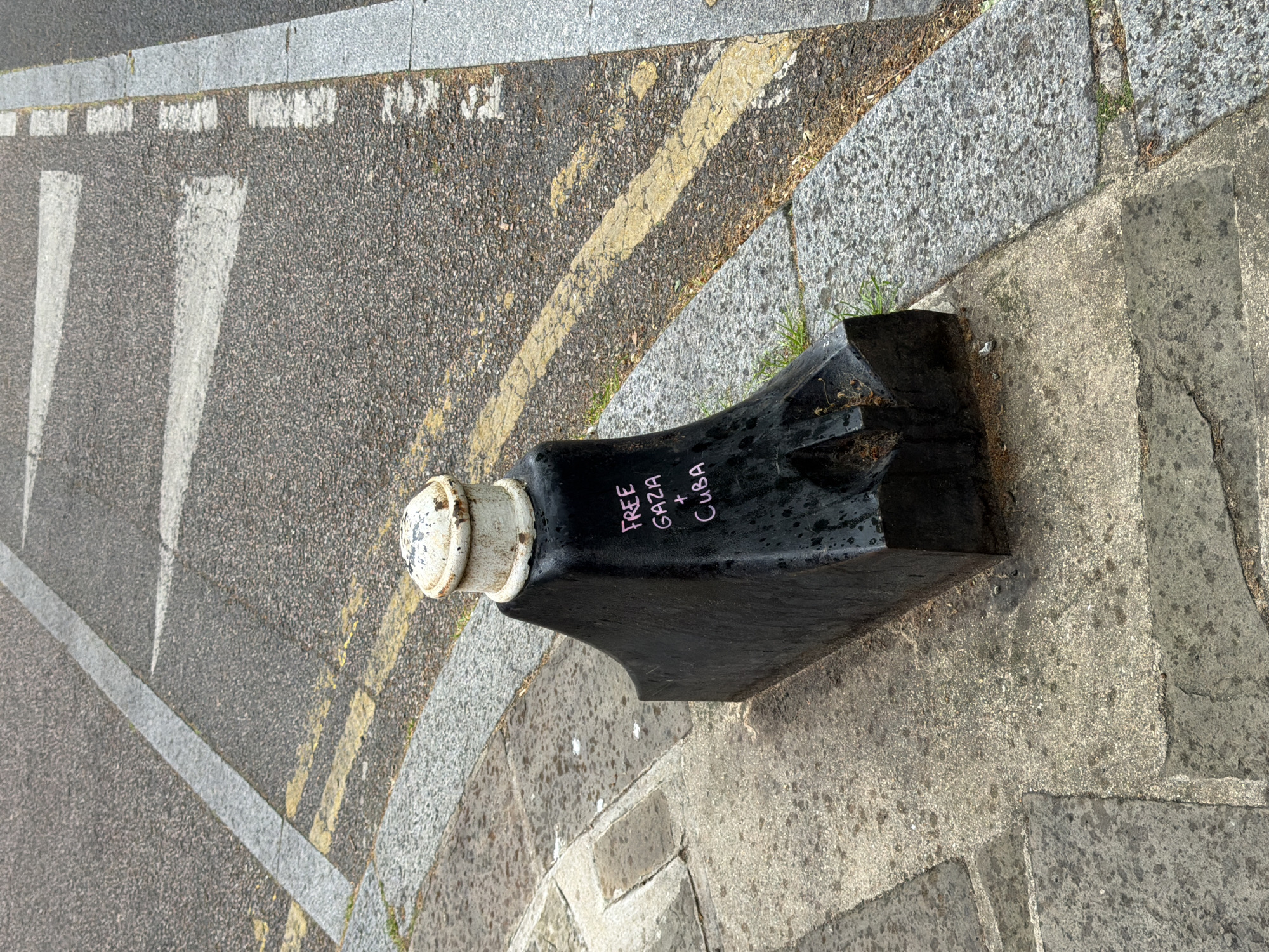 The image shows a black bollard on a street corner in London, United Kingdom. The bollard is cylindrical at the top with a white cap and widens towards the base, which is set into a paved area. Graffiti in pink lettering reading "FREE GAZA + CUBA" is visible on the side of the bollard. To the left of the bollard is a road surface with painted white pedestrian crossing stripes and yellow double lines. The area surrounding the bollard consists of cobblestones and paving stones.