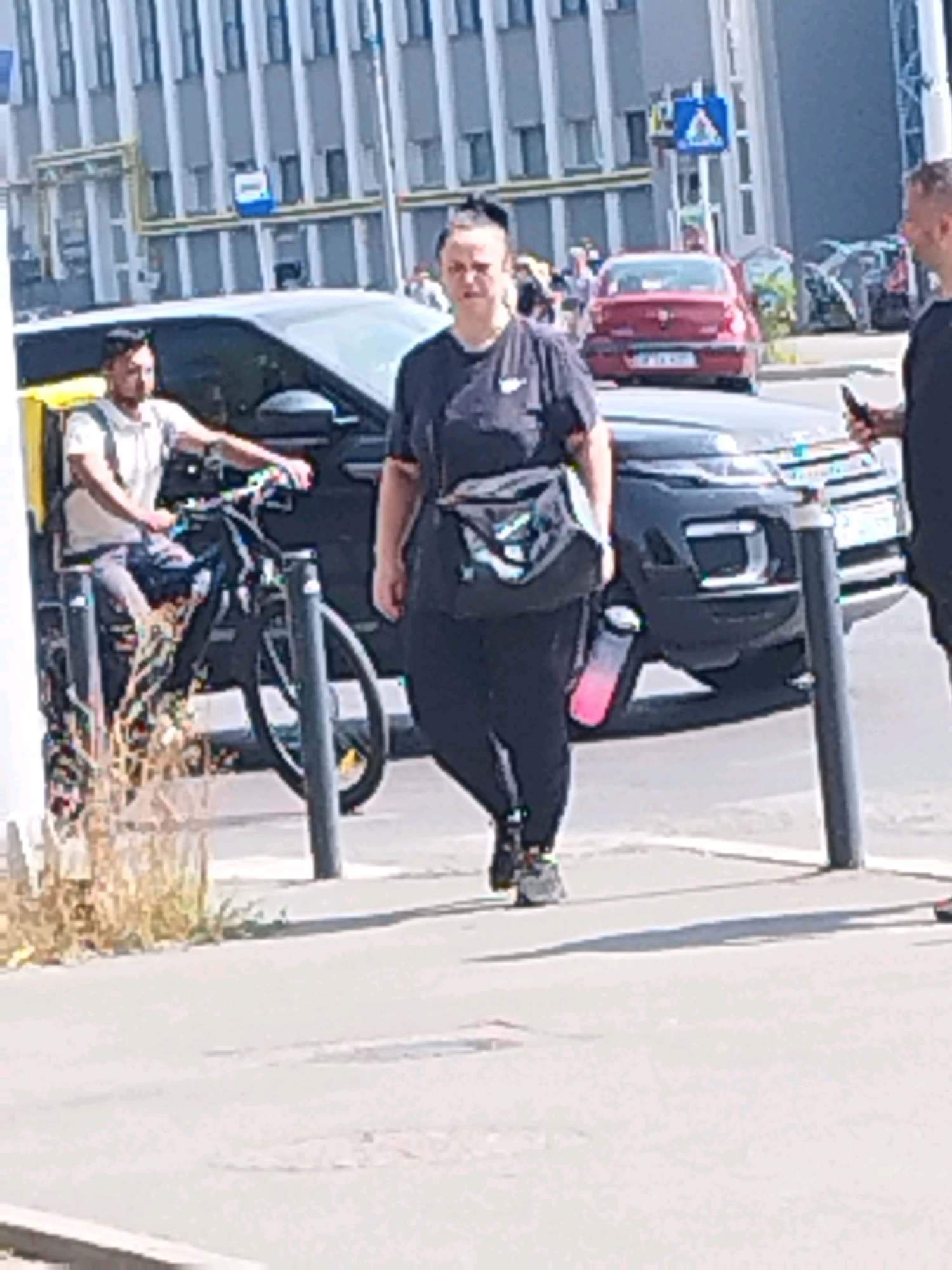 A woman with her hair pulled back in a bun is walking on a sidewalk next to a street in Bucharest, Romania. She is wearing a dark t-shirt and dark leggings. A black crossbody bag is slung across her chest, and a pink water bottle is attached to it. She is wearing dark athletic shoes. To her left, a man is sitting on a bicycle. He is wearing a light-colored polo shirt and dark pants. The bicycle has a yellow delivery box attached to the back. Behind the man and the woman are several vehicles, including a dark SUV and a red sedan. In the background, there is a modern building with a repeating pattern of windows and light-colored horizontal bands. Traffic signs are visible on poles along the street. The scene appears to be in a commercial or urban area.