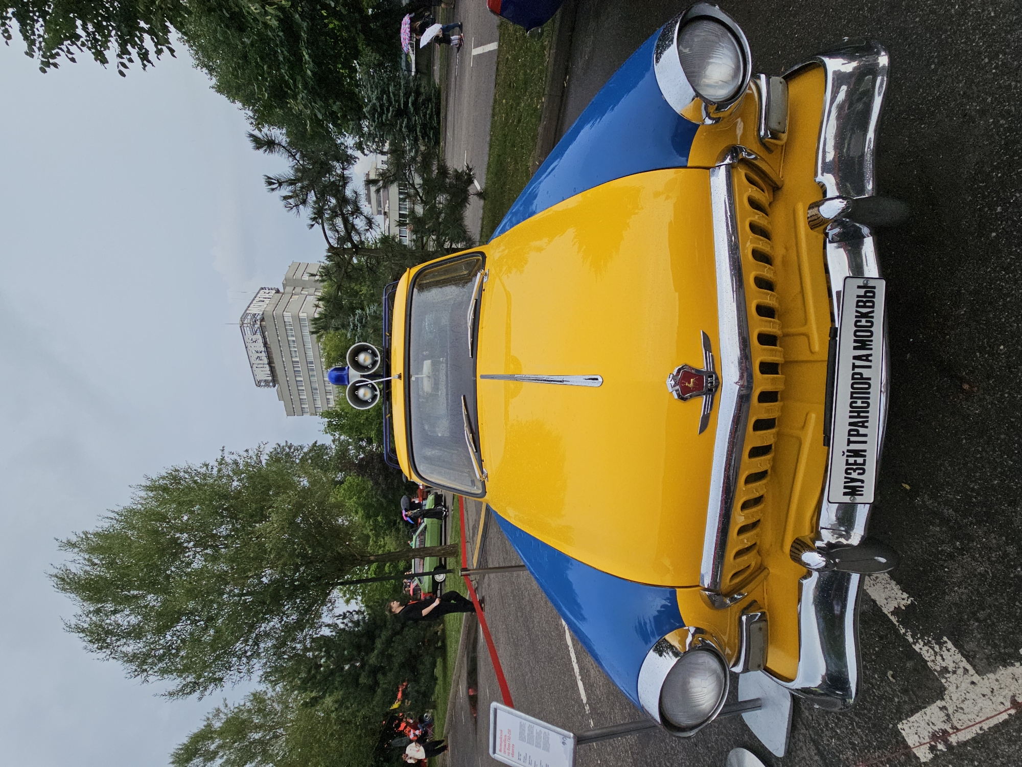 In a parking lot under an overcast sky, a vintage yellow and blue GAZ-21 Volga car is displayed, its front facing the camera. The car features chrome accents on its grille, headlights, and bumper. Mounted on the roof are two blue flashing lights. A license plate on the front bumper reads "МУЗЕЙ ТРАНСПОРТА МОСКВЫ" (Moscow Transport Museum). A red velvet rope barrier runs in front of the car, and a white information sign stands to its left. In the background, several trees, a modern multi-story building, and a few people are visible, some holding umbrellas. The ground is paved and appears wet, with some white painted markings.
