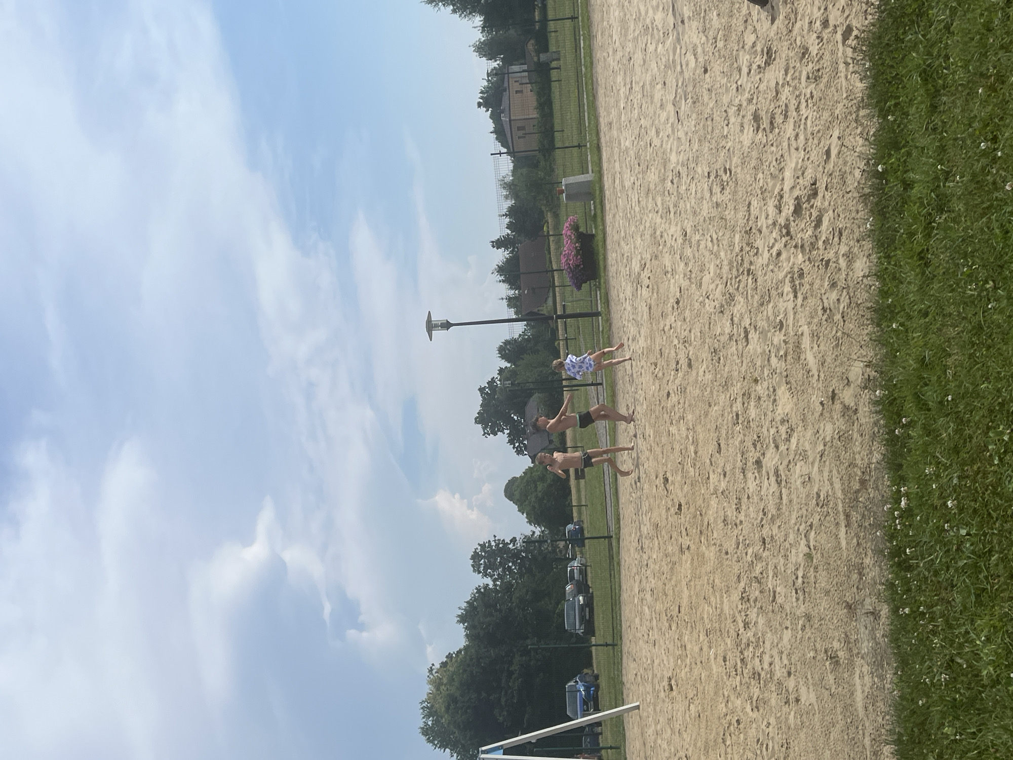 Three young males are lying on a sandy beach volleyball court under a bright, cloudy sky, suggesting it's daytime. The sun is out, but clouds are present, indicating possibly mixed weather. The men are dressed in swim trunks and appear relaxed. One is wearing patterned swim trunks, while the other two have solid colored ones. The court is bordered by a grassy area with some small white flowers. In the background, there are trees, a fence, a lamppost, a planter with pink flowers, and some buildings. A few cars are parked in the distance. The scene suggests a recreational activity on a warm day. No visible text is present in the image.