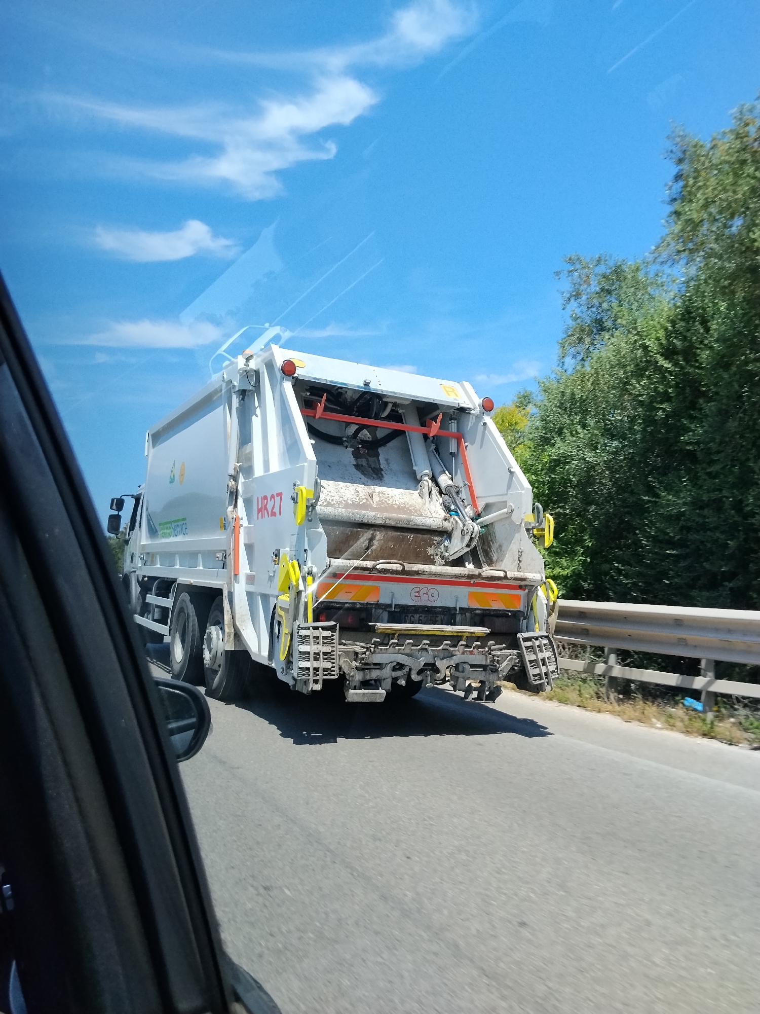 A white refuse compactor truck, identified by "HR27" in red lettering and "SAM Service" branding, is shown from its rear-right perspective, in motion on an asphalt road. The truck's rear compaction mechanism appears heavily soiled, with hydraulic components and yellow attachment points visible. A partial license plate starting "CC 6..." and the word "ECO" are discernible near the lower rear bumper, which also features reflective amber strips. Metal steps are suspended beneath the bumper. The image is captured from the interior of another vehicle, evidenced by a dark window frame and a side mirror on the left. To the right of the road, a metal guardrail separates the highway from dense green foliage. The sky above is clear blue with sparse white cirrus clouds and faint aircraft contrails. Bright daylight conditions are indicated by a distinct shadow cast by the truck onto the road. The scene is located in Molfetta, Italy.