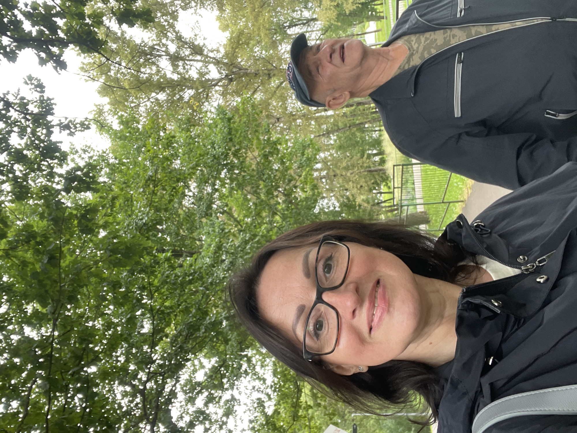The image shows a selfie taken by a woman in her 40s with dark hair and glasses, smiling at the camera. She is wearing a black jacket over a white shirt. To her right, partially visible, is an older man wearing a dark baseball cap, a dark jacket, and a camouflage t-shirt. They are outdoors in a park-like setting with trees in the background. The lighting suggests it is daytime. The presence of a metal fence and paved pathway indicates a public park or recreational area. The overall atmosphere is casual and relaxed. Without further context or landmarks, it is impossible to definitively determine the exact location within the city of Kirov, Russia, though the environment suggests a common urban green space.