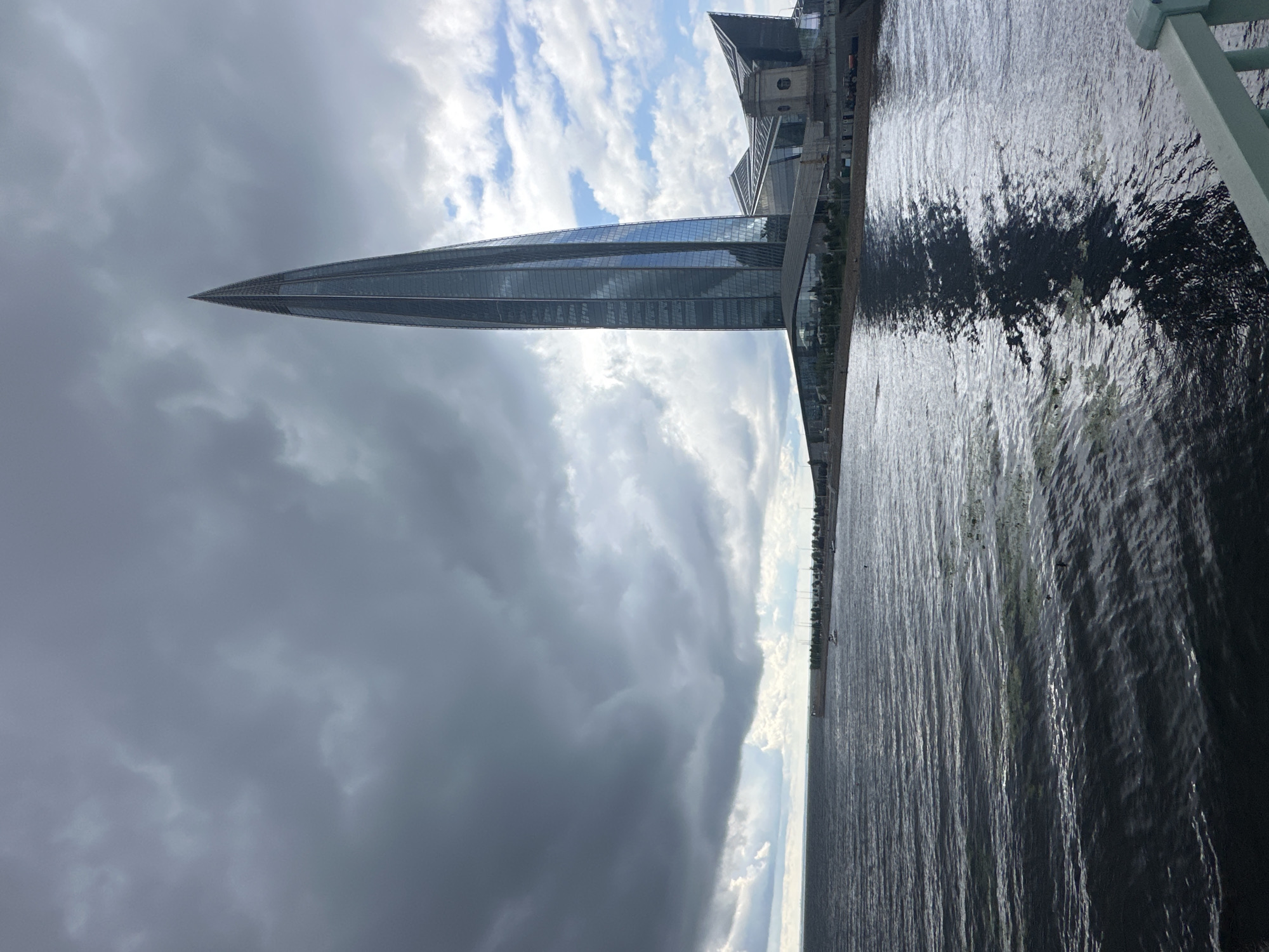 The image captures a view of the Lakhta Center in Saint Petersburg, Russia, from across a body of water. A very tall, slender skyscraper with a pointed top dominates the right side of the frame, its glass facade reflecting the sky and surrounding architecture. The skyscraper is flanked by lower, angular buildings that form a complex.
The body of water in the foreground is choppy, with ripples and reflections of the sky and buildings. Along the far bank, a line of trees or buildings can be seen, with a few small boats on the water. The sky is a mix of dark, dramatic storm clouds and patches of lighter, partly cloudy sky, suggesting a dynamic weather condition. The overall scene is a juxtaposition of modern architecture against a natural and atmospheric environment.