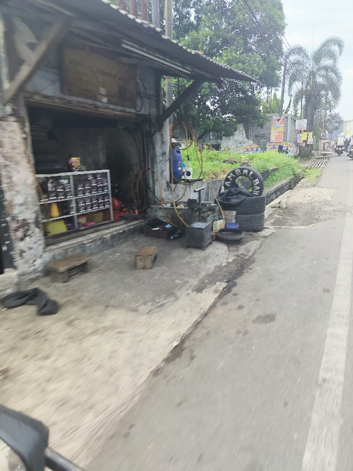 The image shows a small, roadside shop in Ngamprah, Indonesia (latitude: -6.859, longitude: 107.499). The shop is located on the side of a street with a paved surface, and a small, open area sits outside. The shop appears to be closed, with an open doorway leading to a storage space or workshop. The storage space has a shelf unit with various items stored on it. Several items are scattered on the ground in front of the shop, including a tire, a blue toolbox, an air compressor, and a wooden crate. The scene is cluttered and appears to be associated with a tire repair or automotive service. No people are visible in the image. Several trees are present on the edge of the street and there are a few buildings visible in the distance along with several other shops.