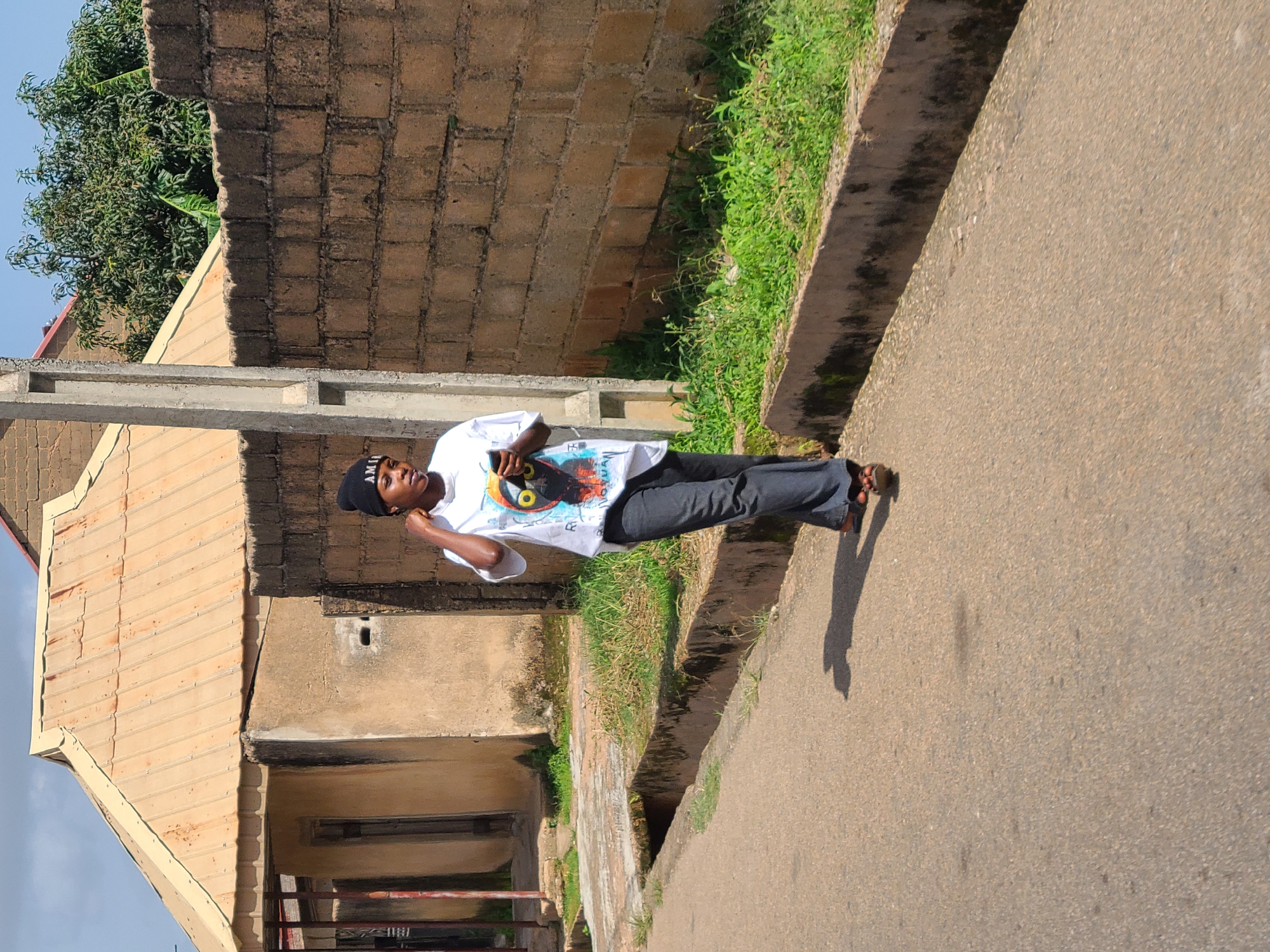 A young person, appearing to be a female, is walking down an asphalt road in Jos, Nigeria, on a sunny day. They are wearing a black beanie, a white t-shirt with a graphic print, and dark denim jeans. The person is holding a mobile phone in their right hand and their left hand is touching their neck. The environment includes a beige corrugated metal roofed building with a rough plastered wall, a section of brown brick wall, a concrete utility pole, and lush green grass growing along a drainage ditch. The road shows some signs of wear and tear. The shadow of the person is cast on the road.