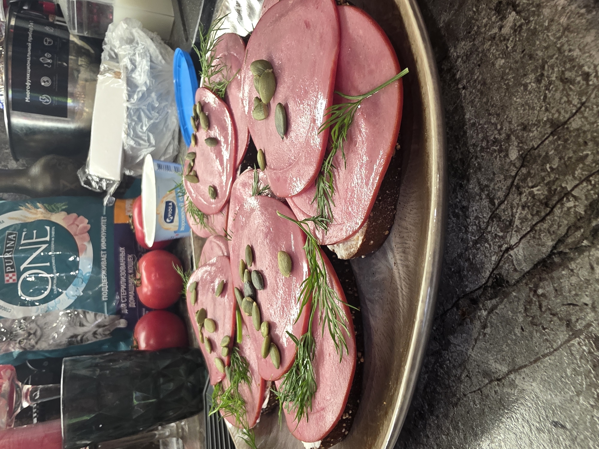 A close-up, eye-level shot showcases several open-faced sandwiches on a round, textured, metallic plate. Each sandwich consists of a dark bread base, a white spread, several slices of pinkish-red processed meat, sprigs of fresh dill, and a scattering of green pumpkin seeds. The sandwiches are arranged in a circular pattern, filling the majority of the plate. In the background, out of focus, are various household items. To the left, a dark, faceted glass is visible. Behind the glass and to the right are two red tomatoes and a container of what appears to be yogurt or cream cheese with a blue lid. Further back, a bag of Purina ONE cat food with a cat image is partially visible. To the right of the cat food bag, a stainless steel appliance, possibly a multi-functional kitchen appliance, is present. The overall environment suggests a domestic setting, likely a kitchen counter with a dark, marbled countertop. The image does not contain any explicit information about a specific location in Saint Petersburg, Russia.