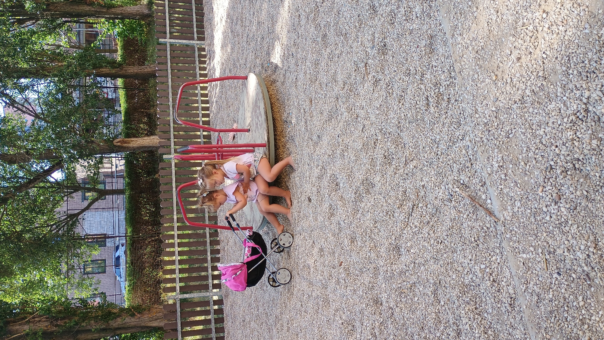 Two young girls are sitting on a circular merry-go-round with a pink and black doll stroller to their left. They are wearing casual summer clothing. The merry-go-round is located on a playground covered in small, light-colored gravel. A wooden fence is visible behind the merry-go-round, with trees and a building in the background. The environment suggests a park or a public play area within a residential or urban setting. The image appears to have been taken during daytime, with sunlight casting shadows on the gravel.