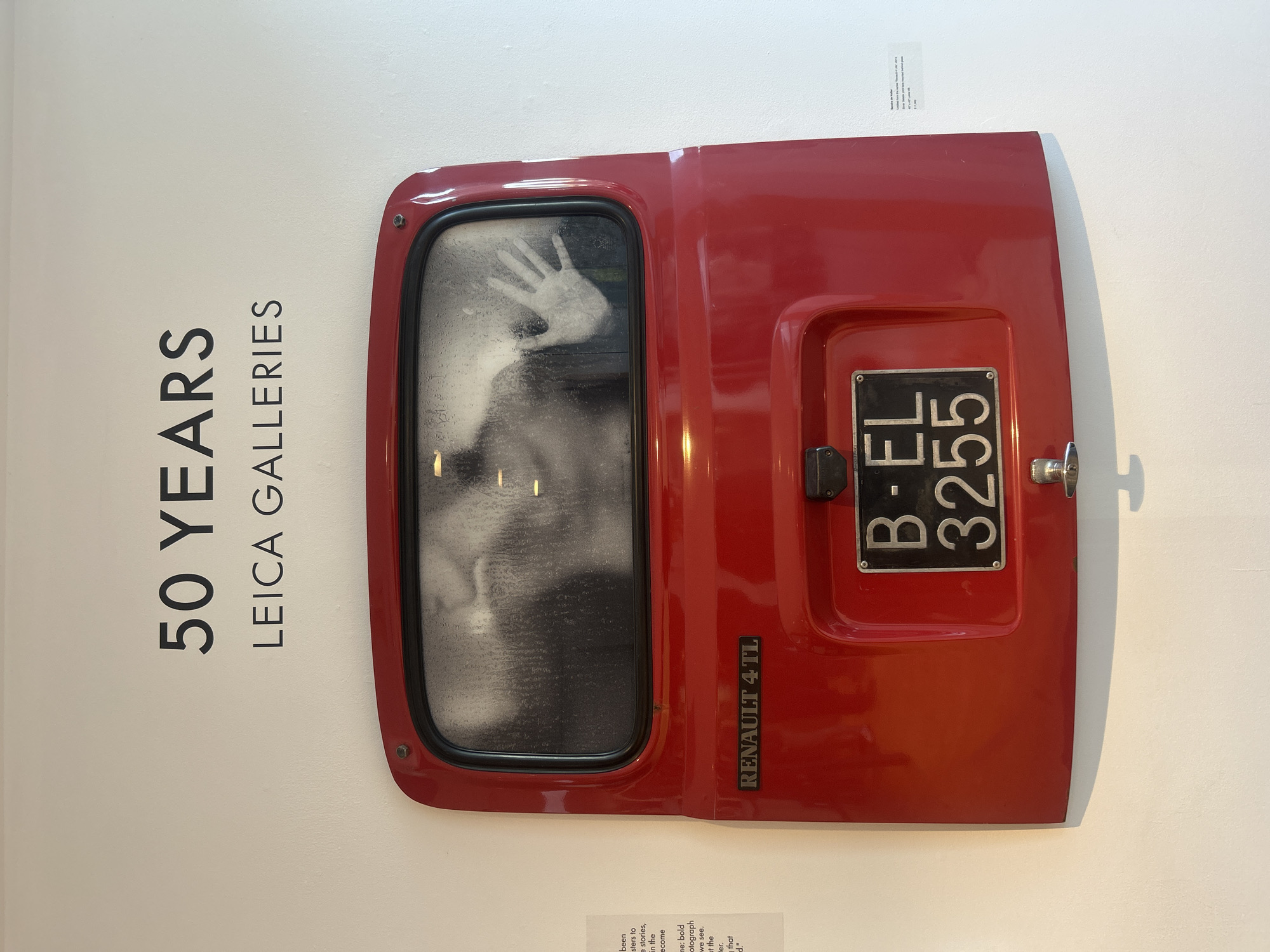 The image displays an artistic installation or exhibit within the Leica Galleries in West Hollywood, USA. The primary subject is a vibrant red car door, identified as a "RENAULT 4 TL" by text embossed on its lower right side. The door is presented vertically against a clean white wall.
Within the car door's window, a stark, blurry monochrome image is visible. The most prominent feature is a ghostly white hand print with splayed fingers, seemingly pressed against the glass from the inside. Behind the hand, a faint, indistinct human form or face can be perceived, creating a sense of mystery or distress. The window's surface appears textured, possibly imitating condensation or frost.
A black license plate bearing the alphanumeric code "B-EL 3255" is attached to the right side of the red door, with a chrome door handle positioned to its right.
To the left of the car door, large, vertically oriented black letters on the white wall announce "50 YEARS LEICA GALLERIES," indicating the nature of the exhibition. Above the car door, a small information card is partially visible, with the title "Sound of a killer" clearly readable, and other smaller text below it. Another information card is partially visible below the car door, containing fragmented text such as "been," "stories," "become," "bold," "photograph," and "we see." The setting is an indoor gallery, characterized by bright, even lighting, highlighting the art piece. The installation evokes a thought-provoking atmosphere, possibly touching on themes of confinement, memory, or human experience.