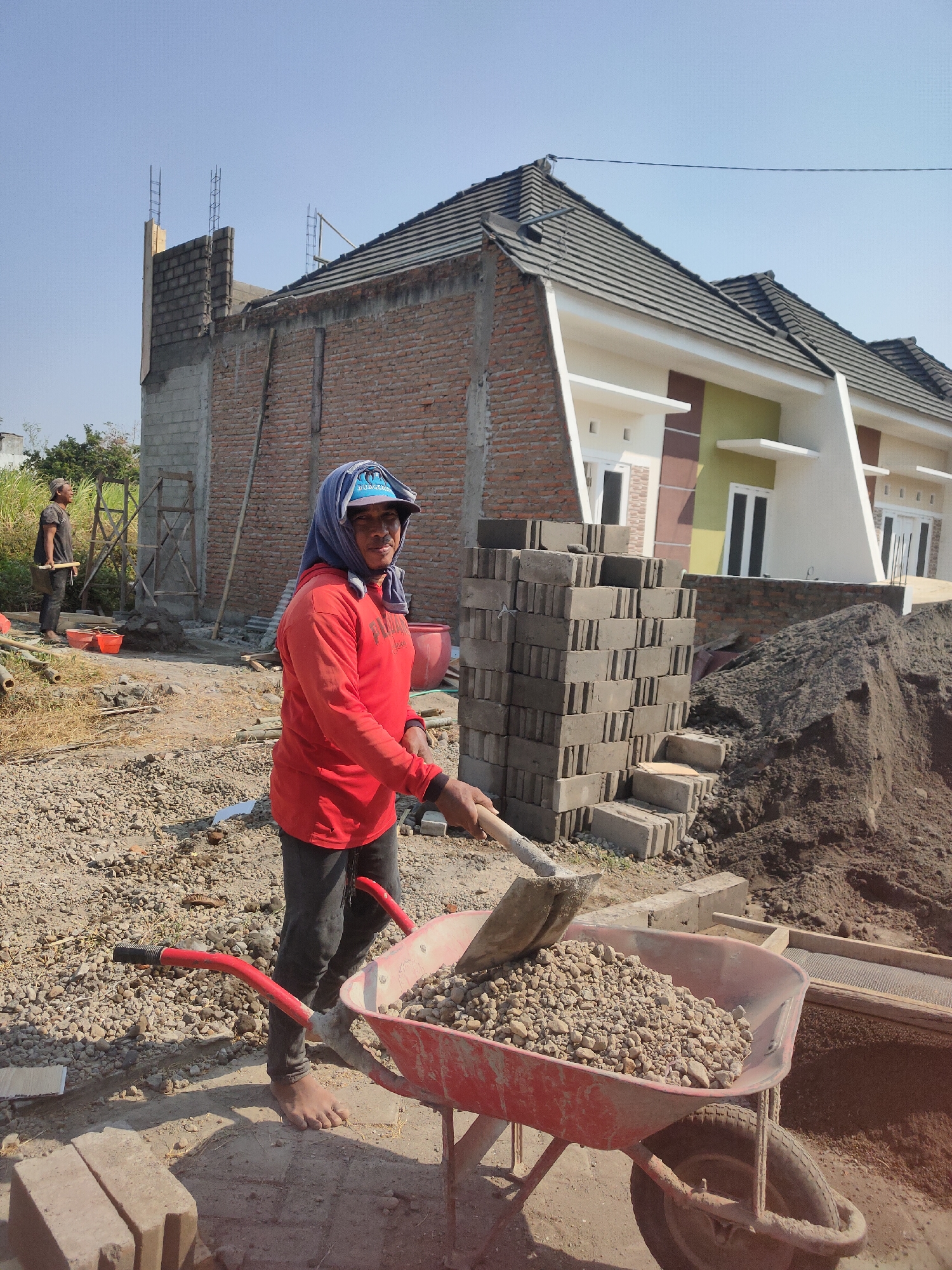In Ngasem, Indonesia, a construction site under a clear blue sky shows two men working. The foreground features a man in a red long-sleeved shirt and dark pants, wearing a blue headscarf and a blue baseball cap. He is barefoot and using a shovel to fill a red wheelbarrow with gravel. The wheelbarrow is positioned on loose gravel and dirt. Behind him, to the left, another man in a dark shirt and pants stands holding a shovel, looking towards the unfinished construction.
The construction area includes partially built brick walls and concrete foundations. A stack of gray bricks is visible to the right of the foreground worker, and a large pile of dark soil is next to it. In the background, a multi-unit residential building with gray tiled roofs, white and green facades, and dark-framed windows is under construction. The scene is illuminated by bright sunlight, casting shadows on the ground.