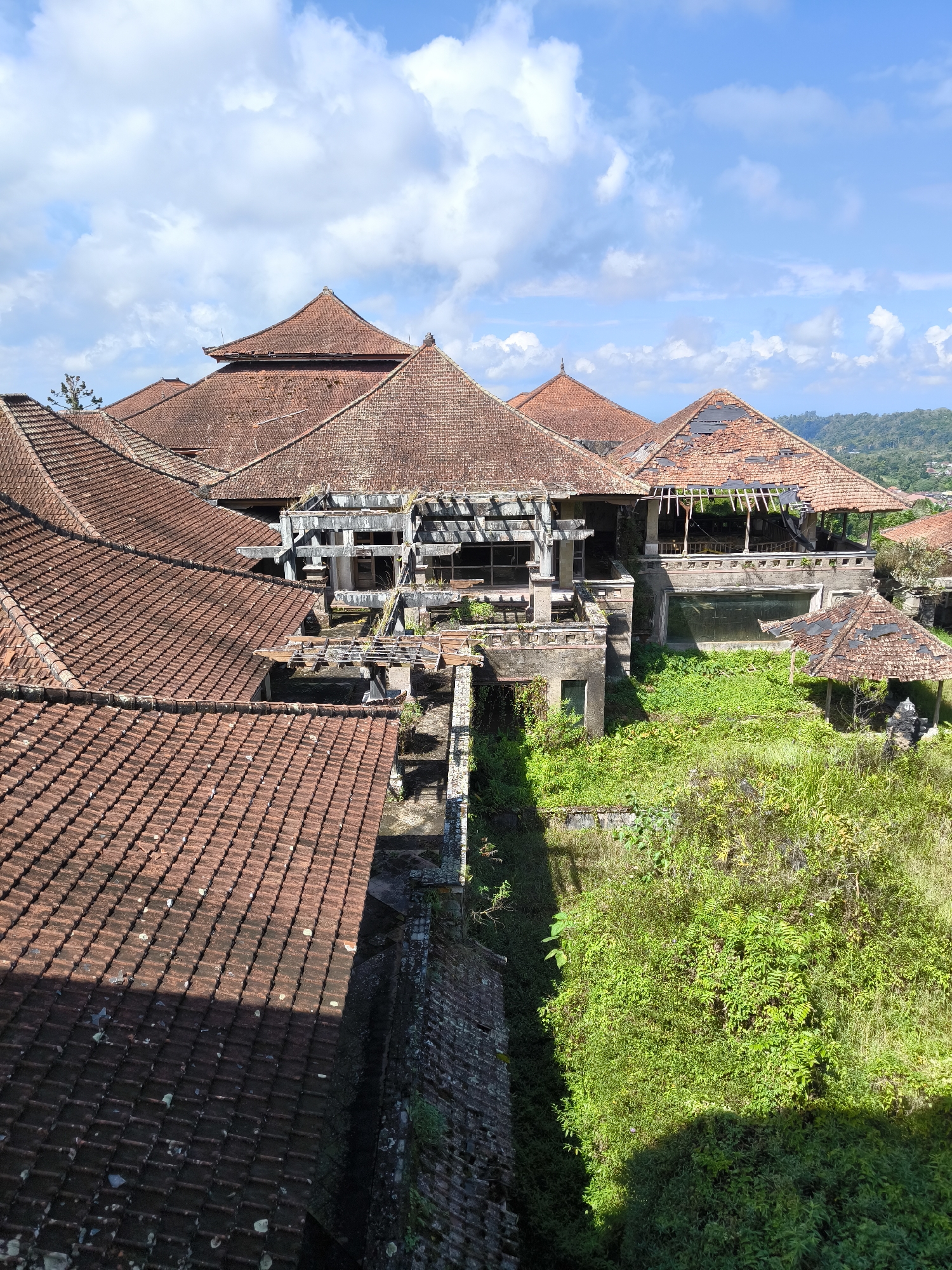 The image depicts an abandoned resort in Baturiti, Indonesia, under a bright blue sky with scattered white clouds. The resort features multiple buildings with traditional terracotta tiled roofs, many of which are in a state of disrepair with missing or broken tiles, revealing the wooden structures beneath. The architecture appears to be Balinese-inspired, with multi-tiered roofs and open-air structures.
The resort is overgrown with lush green vegetation, with trees, bushes, and vines encroaching on the buildings. The once manicured grounds are now wild and untamed. The buildings themselves show signs of decay, with crumbling concrete, broken windows, and peeling paint. Some areas of the resort seem to have been designed with courtyards or water features, now filled with foliage.
The overall impression is one of abandonment and nature reclaiming the man-made structures. The scene is still and silent, with no people visible. The surrounding landscape suggests a hilly or mountainous terrain, with distant hills covered in greenery. The image captures a sense of lost grandeur and the passage of time.