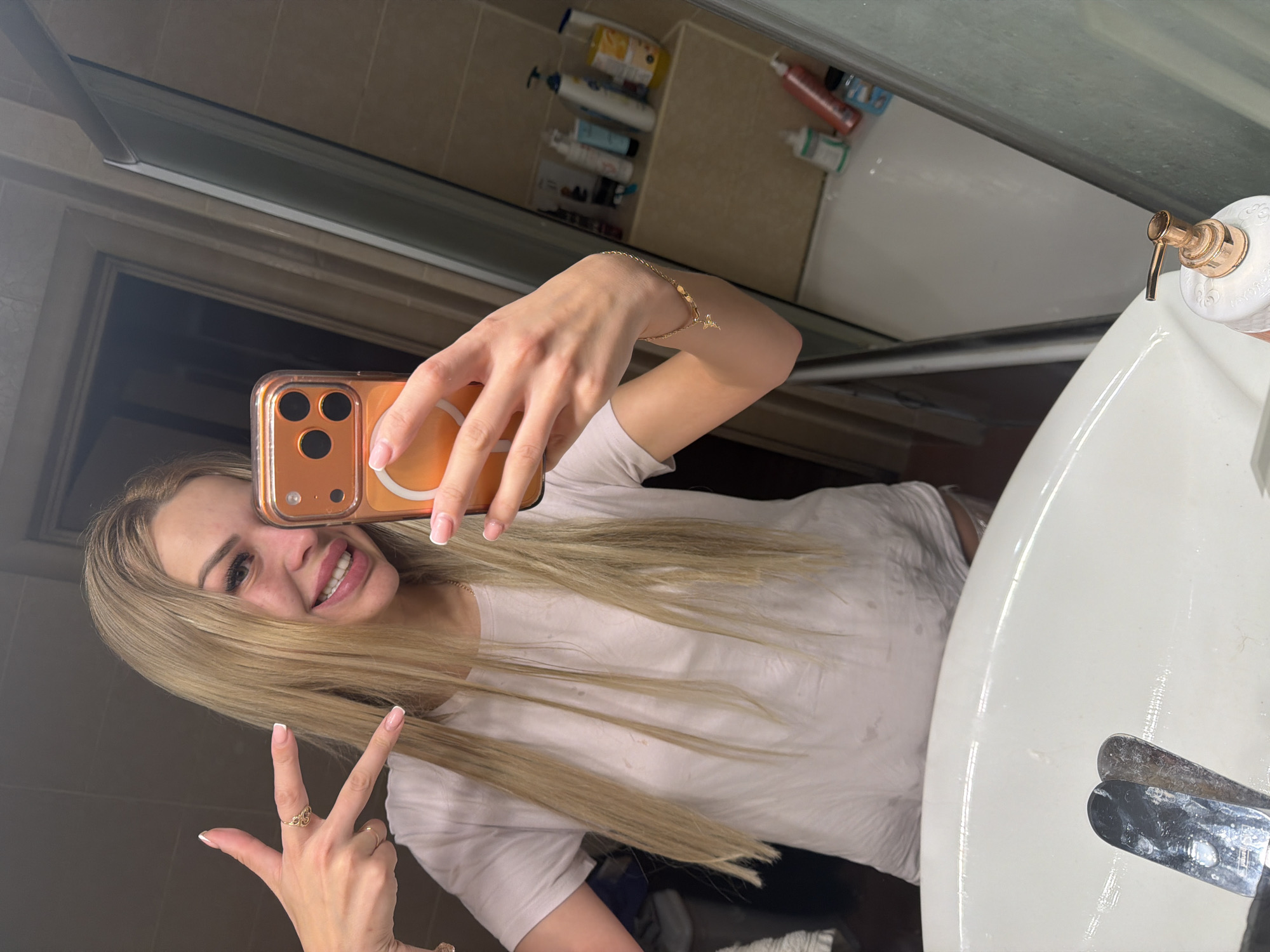 A young blonde woman is taking a mirror selfie in a bathroom. She is wearing a white t-shirt and has her hair down. She is holding a phone in her right hand and making a peace sign with her left hand. The bathroom appears to be well-lit with beige tiles and various toiletries visible on shelves. There is a sink in the foreground and a bathtub in the background. The overall scene is casual and personal.