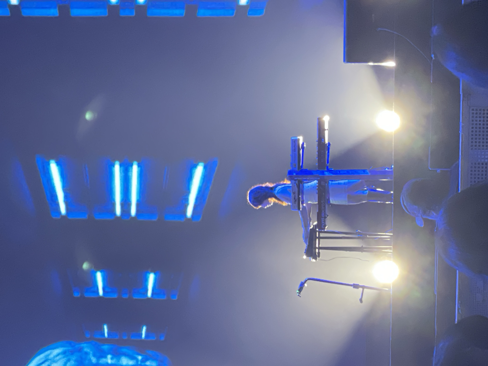 The image shows a woman playing a keyboard on a stage, illuminated by bright white spotlights. The stage is equipped with lighting fixtures emitting a blue glow. A microphone stand is visible near the keyboard. Stage equipment and rigging are present. The exact location in Salembaran, Indonesia cannot be determined from the image.