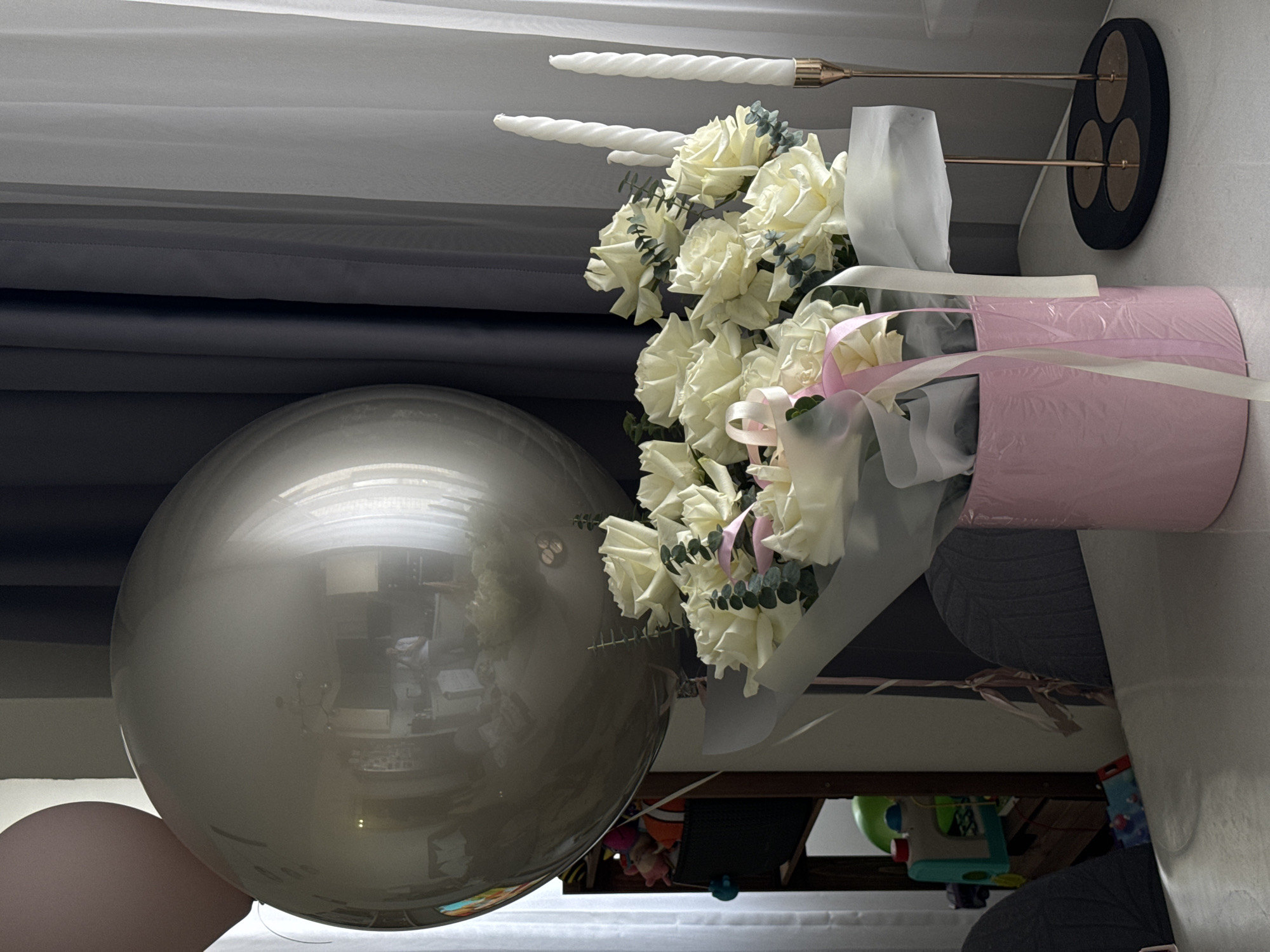 The image captures a festive indoor scene, likely arranged for a celebration. On the left side of the frame, a prominent, large, spherical grey or silver balloon reflects the surrounding room, showing indistinct shapes of furniture and possibly a light fixture. A smaller, muted pink or beige balloon is partially visible below and to the left of the larger one.
To the right, a beautiful bouquet of fresh white roses is presented, interspersed with green eucalyptus foliage. The bouquet is stylishly wrapped in a pink cylindrical container or material, accented with delicate pink and white ribbons. Above the bouquet, in the upper right, a modern candle holder stands with a dark circular base and two gold-toned metallic arms, each holding an unlit, white, twisted taper candle.
The background on the upper left features soft light filtering through sheer white and opaque grey curtains, suggesting a window. Below the floral arrangement, in the lower right, some indistinct household items or toys are partially visible, hinting at a lived-in space. The overall arrangement of balloons, flowers, and candles evokes a celebratory mood, typical of events like birthdays or anniversaries. No people are visible, and there is no discernible text. The scene depicts a well-decorated room, likely within a private residence in Odintsovo, Russia, during daytime hours.