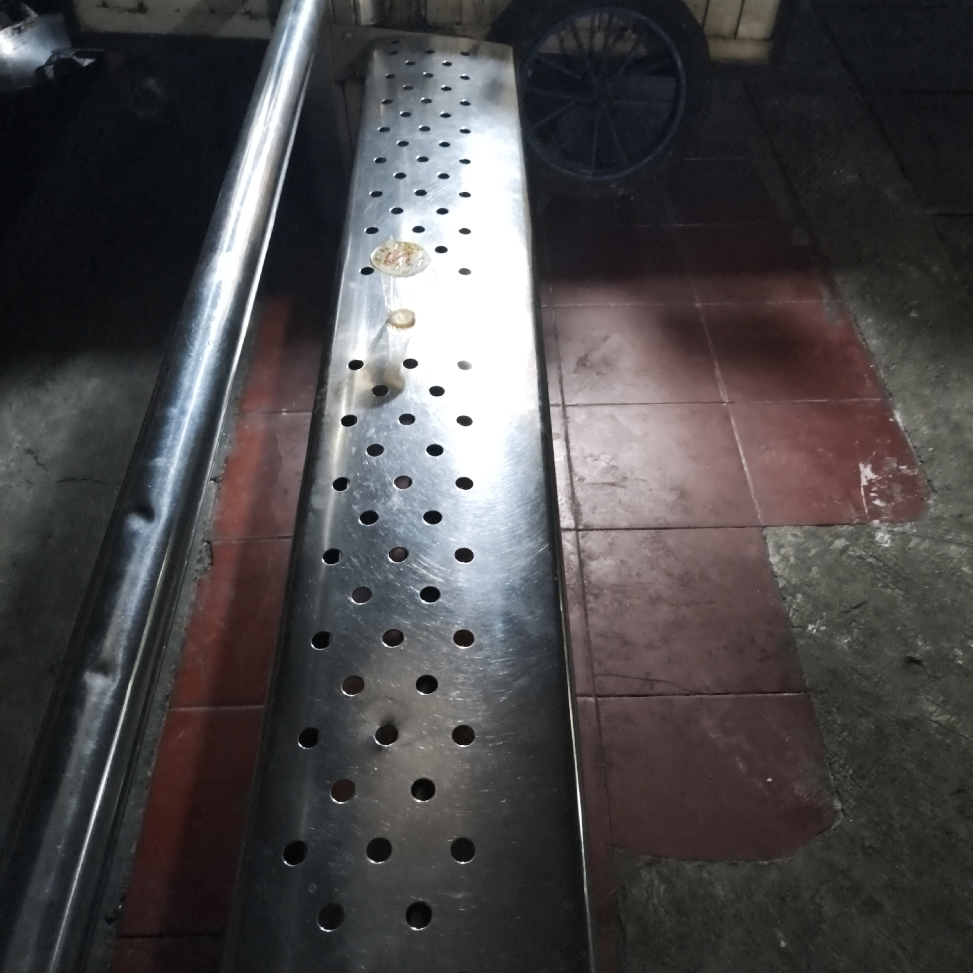 In South Jakarta, Indonesia, an image captured from a low angle depicts a metal ramp with numerous circular perforations, running parallel to a polished metal pole. The ramp is positioned on a tiled floor, with visible grout lines indicating red tiles adjacent to a dark, possibly concrete, surface. In the background, a black wheel with spokes is partially visible. The lighting suggests an indoor or covered outdoor setting, possibly a garage or workshop, with reflections on the metal surfaces. There are no people or animals present in the image, and no discernible activity is occurring. The scene appears static and devoid of any specific narrative.