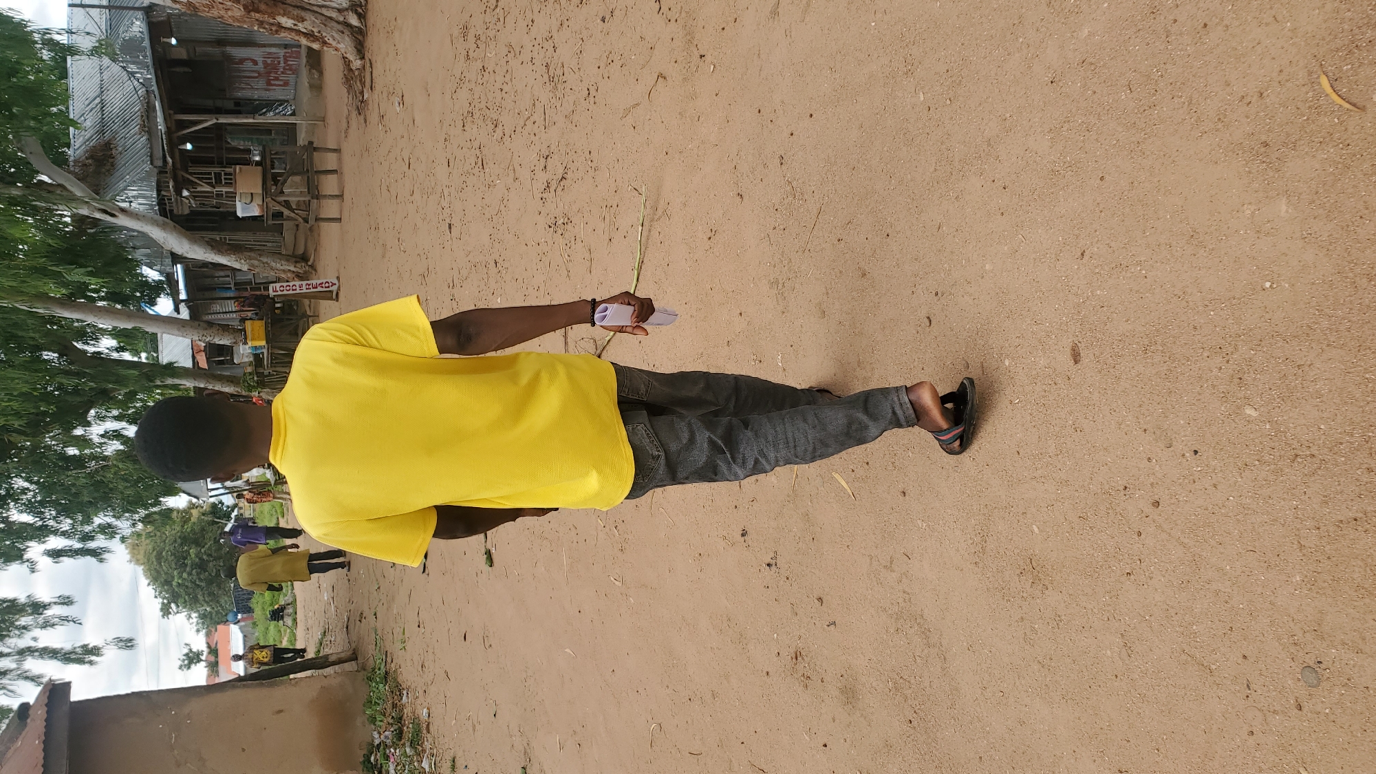 A young Black male is walking away from the camera on a dirt path in what appears to be a village or semi-urban area. He is wearing a bright yellow t-shirt and dark grey, possibly denim, jeans. His left foot is visible, wearing a dark sandal with red and blue straps. In his right hand, he holds a rolled-up piece of white paper. Behind him, several other individuals are visible in the distance, also walking along the path. To the right of the path are several small, open-air structures with corrugated metal roofs, appearing to be market stalls or shops. Trees with large, light-colored trunks and green foliage line the path and surround the structures. The ground is a sandy, reddish-brown color, with scattered debris and small stones. The sky is overcast. The overall impression is a snapshot of daily life in a dusty, outdoor environment.