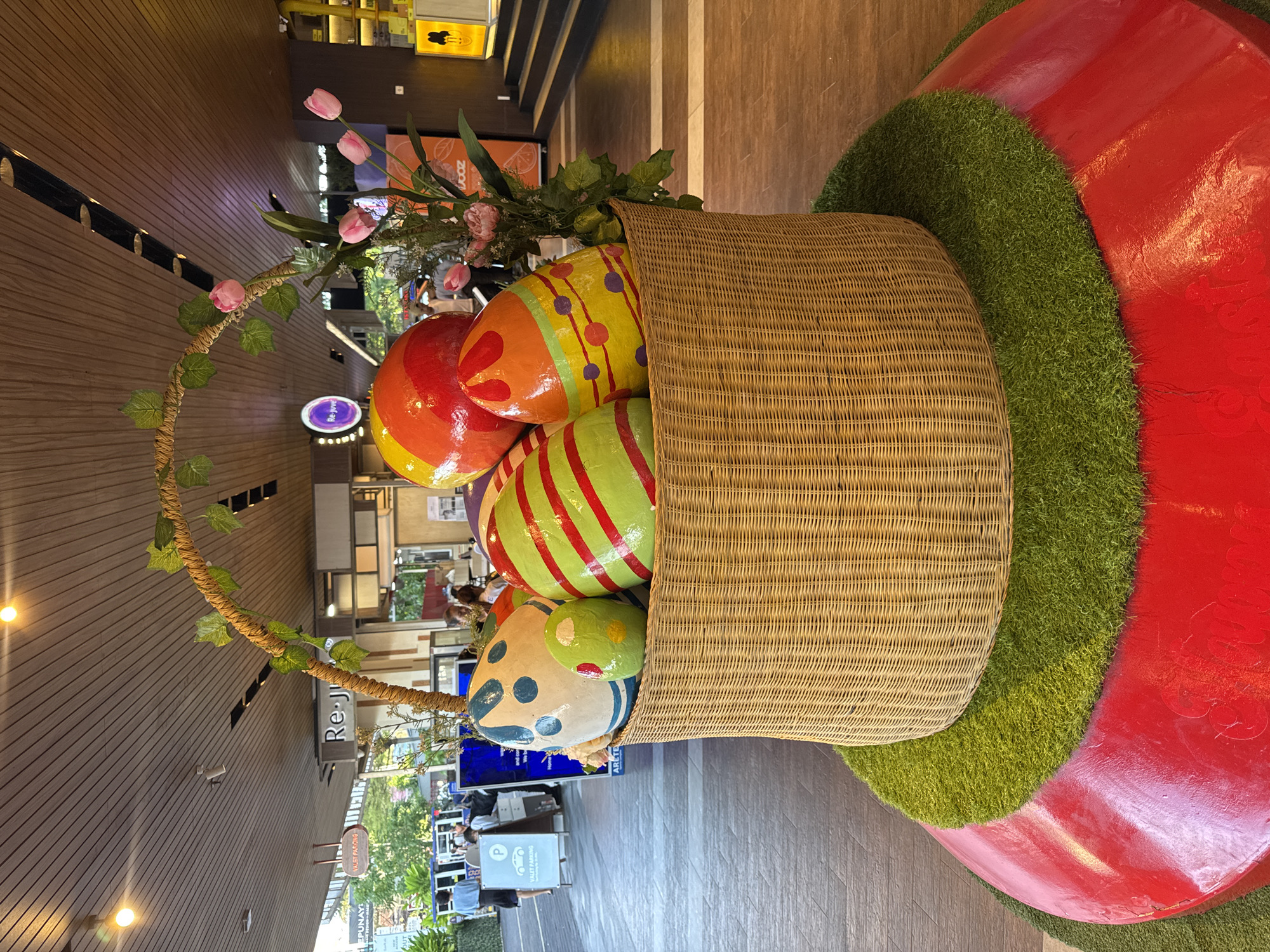 This image captures a vibrant Easter display set up in an indoor commercial space, likely a shopping mall, in Kuta Selatan, Indonesia. The central focus is a large, light brown wicker basket overflowing with several oversized, brightly colored Easter eggs.
The eggs are distinct, each painted with festive patterns: one is predominantly red and orange with yellow accents, another yellow with purple polka dots and red stripes, a third green with bold red stripes, and a fourth blue with white and green polka dots. The basket features a decorative handle wrapped in a vine adorned with artificial green leaves and small pink flowers. A bouquet of artificial pink tulips and other blossoms, along with lush green foliage, is placed near the eggs within the basket.
The basket rests on a patch of artificial green grass, which in turn is situated on a large, glossy red, curved base. The background reveals modern architectural elements, including warm-toned wooden paneling on walls and ceilings, and various shops or stalls. Blurry figures of people can be seen moving in the background, indicating a public and active environment. Visible signs include "Re-J" on a purple sign and a "Valet Parking" sign with a prominent 'P' symbol. The indoor lighting suggests a well-lit environment, possibly daytime or evening. The display is a cheerful, holiday-themed decoration, likely intended to enhance the ambiance for shoppers.