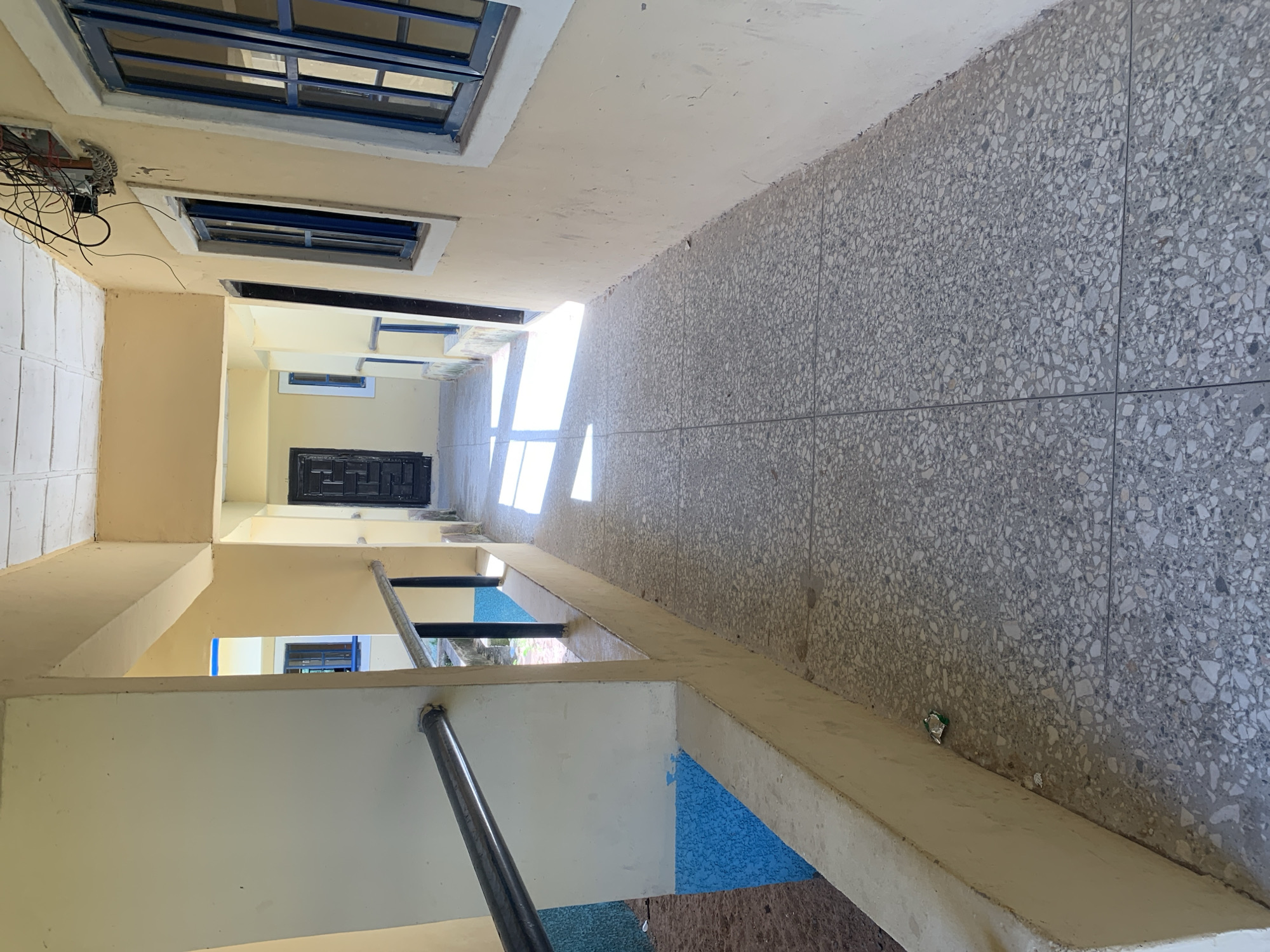 The image depicts an outdoor corridor or walkway in Jalingo, Nigeria. The corridor has a tiled floor with a speckled pattern of gray and white. The walls are painted a pale yellow, and there is a concrete railing with a metal bar running along the left side. On the right side of the corridor, there are several windows with blue frames and dark bars. At the end of the corridor, a black door is visible, and sunlight streams in from an opening on the left, creating a shadow pattern on the floor. There are no people visible in the image. The environment appears to be a residential or institutional building with simple architecture.