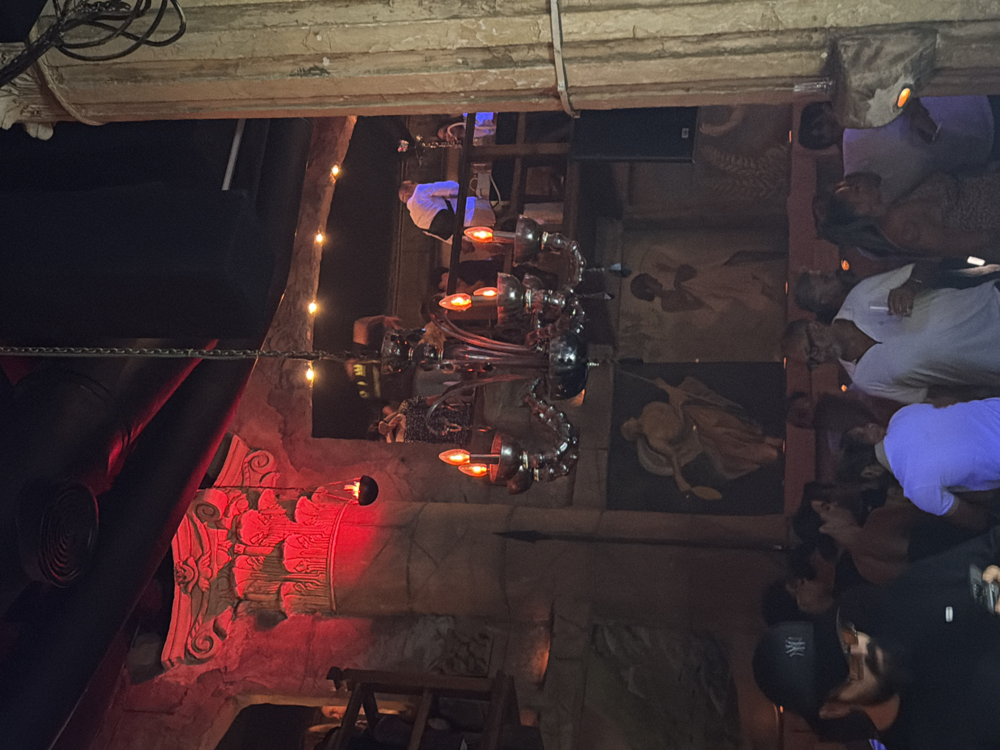 This image depicts the interior of a nightclub or bar, possibly located in Sharm El Sheikh, Egypt, characterized by its opulent and classical-inspired decor. The scene is dimly lit, with red and warm yellow lighting creating an atmospheric ambiance. Prominently featured is a large, ornate chandelier with multiple candle-like lights, hanging from a chain. To the left, a grand, carved stone column or architectural element is illuminated with red light, resembling ancient Egyptian or Roman architectural styles. The background reveals a mezzanine level where several people are visible, some standing and interacting. One individual in a white shirt is leaning over a railing. On the lower level, a crowd of people are gathered, some engaged in conversation and others looking at their phones. The overall aesthetic suggests a sophisticated entertainment venue.