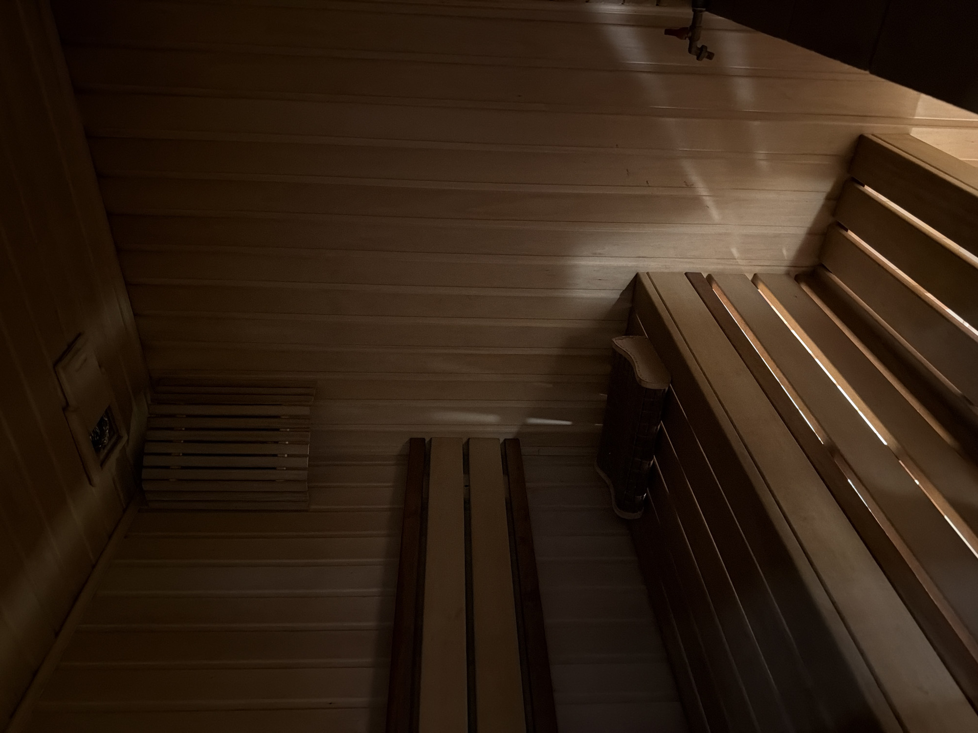 An interior view of a wooden sauna shows a bench with a book on it, illuminated by natural light filtering through the slats of the bench. The walls and ceiling are also constructed of wood. The scene is dimly lit, creating a warm and relaxing atmosphere. The location is identified as the city of Qarağandı city administration, Kazakhstan.