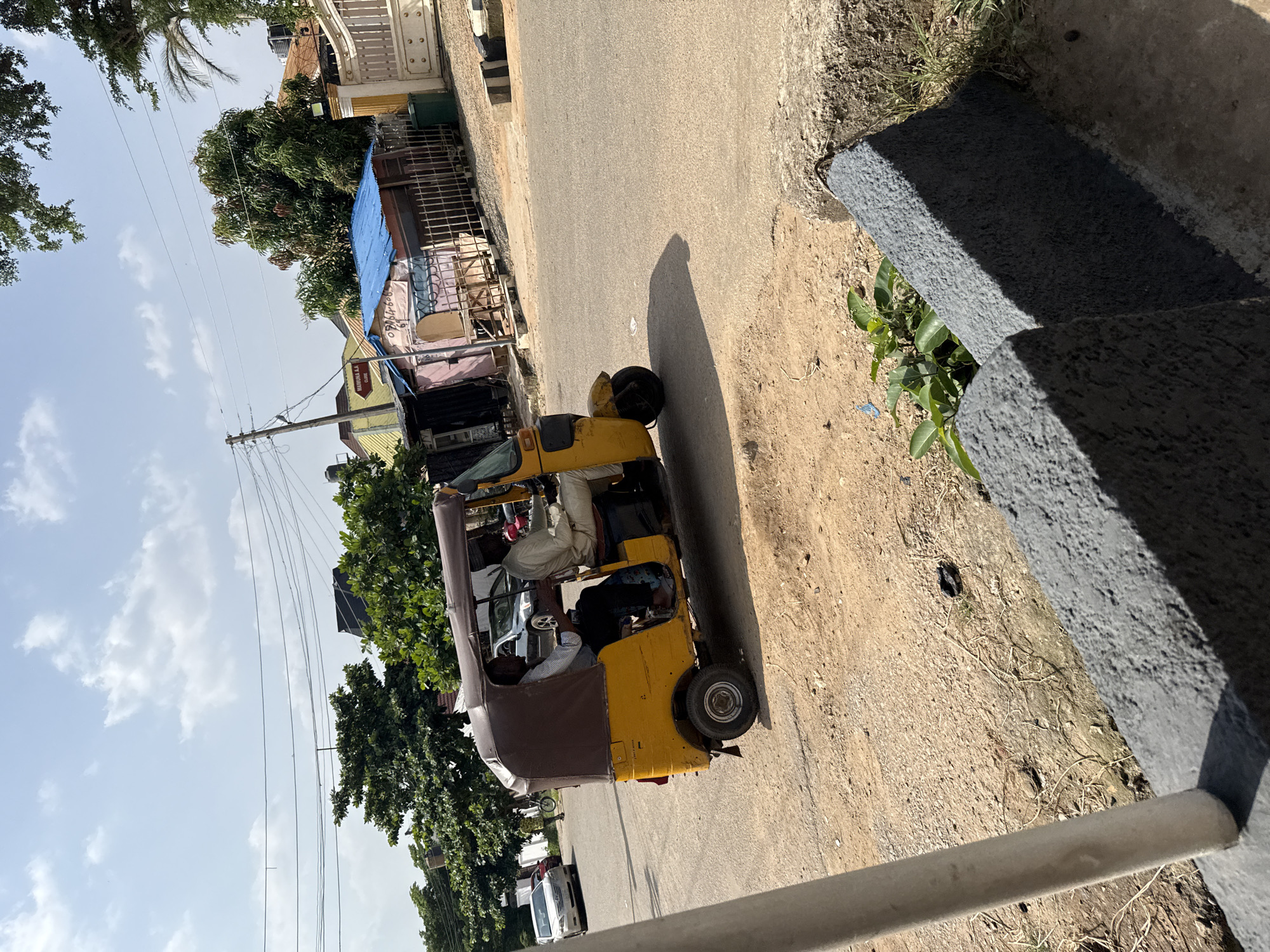 The image depicts a street scene in Abuja, Nigeria, likely during the daytime due to the bright sunlight casting long shadows. The main subject is a yellow and brown auto-rickshaw, also known as a "Keke," parked on the side of an asphalt road. The auto-rickshaw appears to be carrying passengers, with one person visible in the driver's seat and at least two others in the passenger area.
The setting is a roadside with a building or shops on the left, featuring a blue tarp covering part of the structure. Trees with lush green foliage are present, suggesting a warm climate. The road itself is a light gray asphalt, with a dirt and gravel shoulder on the right. A concrete structure, possibly a wall or raised platform, runs along the right side of the image, casting a distinct shadow.
Visible text on a sign above one of the shops reads "AKUGUNA A.J GLOBAL." The overall impression is one of everyday life and transportation in an urban or semi-urban Nigerian environment. The sky is bright blue with scattered white clouds, indicating fair weather.