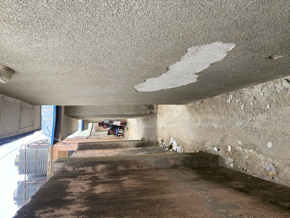 An image depicts a narrow unpaved alley in the city of Katcha, Nigeria, extending forward between two multi-story buildings under a hazy sky. The left wall features weathered reddish-brown rendering, while the right wall is light grey textured stucco with large sections of render peeled away, revealing a lighter subsurface. The unpaved ground consists of dirt and scattered litter, with a slightly raised central path of compacted earth or worn concrete. Approximately five individuals are gathered in the mid-ground of the alley: a shirtless male is seated on a white stool, surrounded by standing adults and a young child in a light-colored outfit. In the background, above the structures, multiple black plastic water storage tanks are visible on a rooftop. Overhanging eaves extend from the buildings above both alley walls. A light fixture is mounted on the upper section of the right wall.