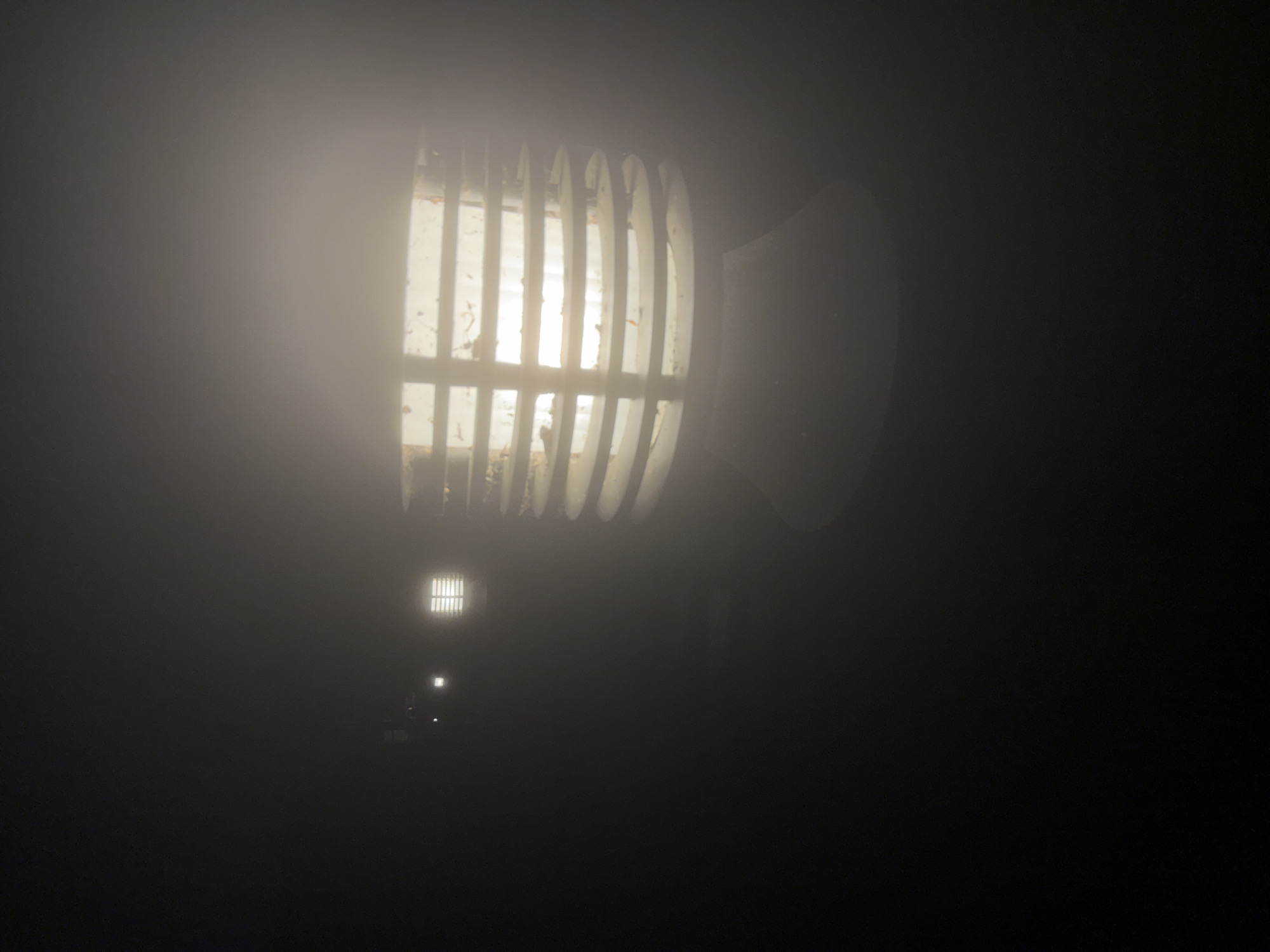 The image shows a foggy night scene. A large, cylindrical outdoor light fixture is the focal point, its metal slats and illuminated interior visible through the fog. The light is mounted on a pole or structure that is not fully visible. Further back, other similar light fixtures recede into the fog. The scene is dark and visibility is extremely limited due to the heavy fog. Without additional information, a precise location in London cannot be determined.