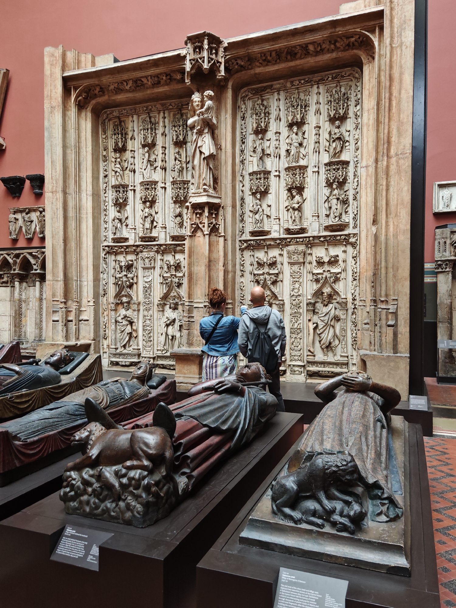 The photograph shows a museum exhibition, possibly in London, United Kingdom, featuring medieval or Renaissance sculptures and architectural elements. Two visitors, a man and a woman, are standing in front of a large ornate stone architectural piece that appears to be an elaborately carved portal or doorway, possibly from a church or palace. The doorway has a central statue of the Virgin Mary holding the baby Jesus. Below, there are four sculpted effigies of deceased individuals lying on tombs or biers, with animals at their feet (lion, dog). Each has a information tag nearby. The walls of the exhibition space are a muted red or terracotta color. The overall atmosphere suggests an exhibition of medieval or Renaissance art and funerary objects.