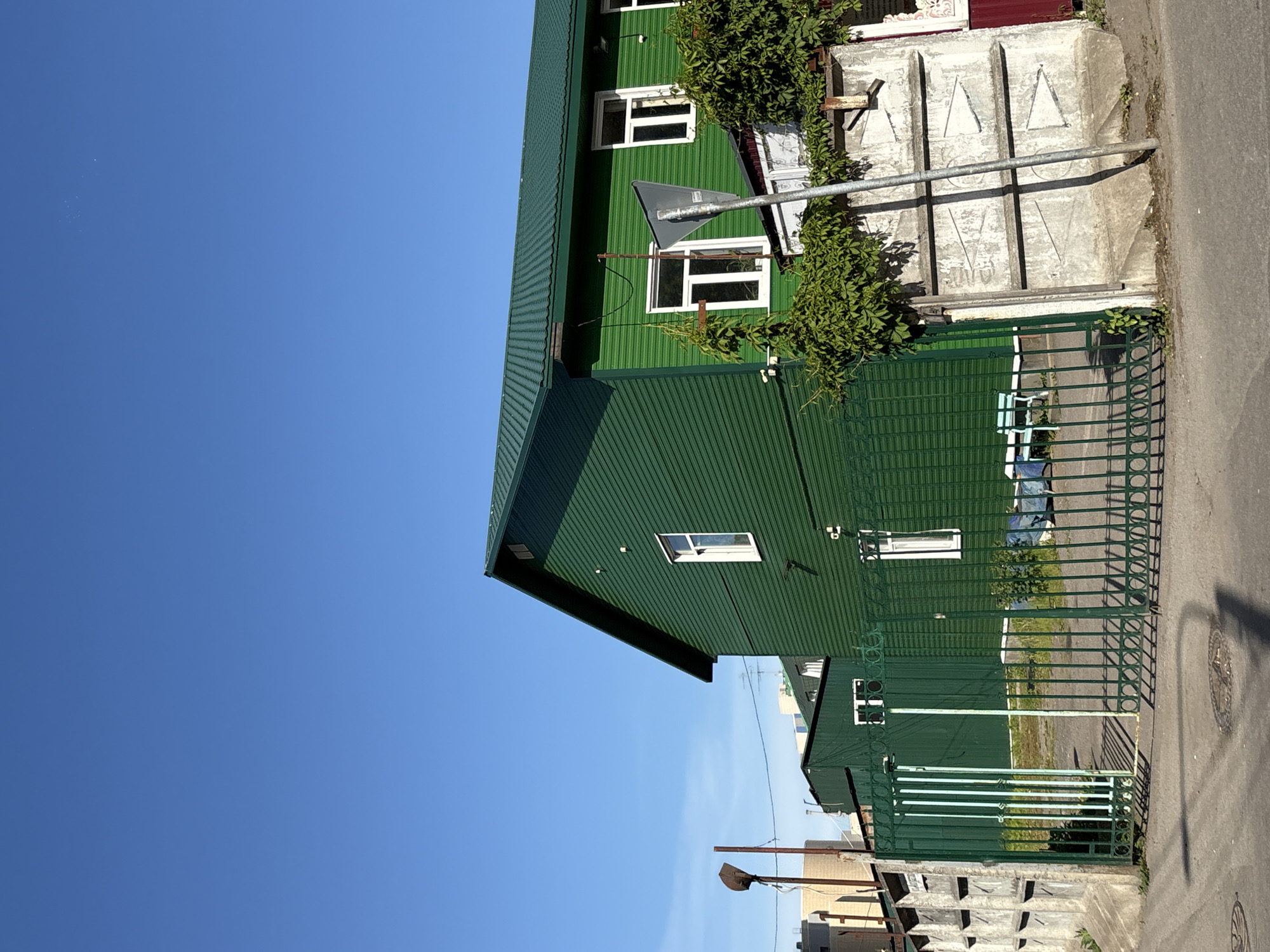 The image displays an outdoor scene dominated by buildings painted in various shades of green, under a clear, bright blue sky. On the right side, a multi-story building features dark green corrugated metal siding and roof, with multiple white-framed windows. Green vines are growing on parts of this building and draped over an adjacent concrete structure.
In the mid-ground, a large, light-grey concrete block structure is visible, featuring embossed or carved triangular patterns on its surface. A gray metal pole stands in front of this concrete structure. Below this, a dark green metal fence, possibly part of a gate, extends across the foreground, separating the buildings from an asphalt road on the left. A lighter green metal gate is partially visible further back behind the fence.
A triangular gray street sign is attached to a pole near the upper building, but its text or symbol is illegible. In the background, hints of other buildings and utility poles with wires can be seen. The asphalt road in the foreground shows a partial view of a manhole cover with a star-like pattern, though any text on it is indistinct.
The scene is illuminated by direct sunlight, casting sharp shadows on the buildings and ground, indicating a clear day, likely late morning or early afternoon. The sky is uniformly blue and cloudless, with a few tiny white specks visible in the upper left area. No people are present. The architectural style and materials, such as corrugated metal siding, are common in some regions of Russia.