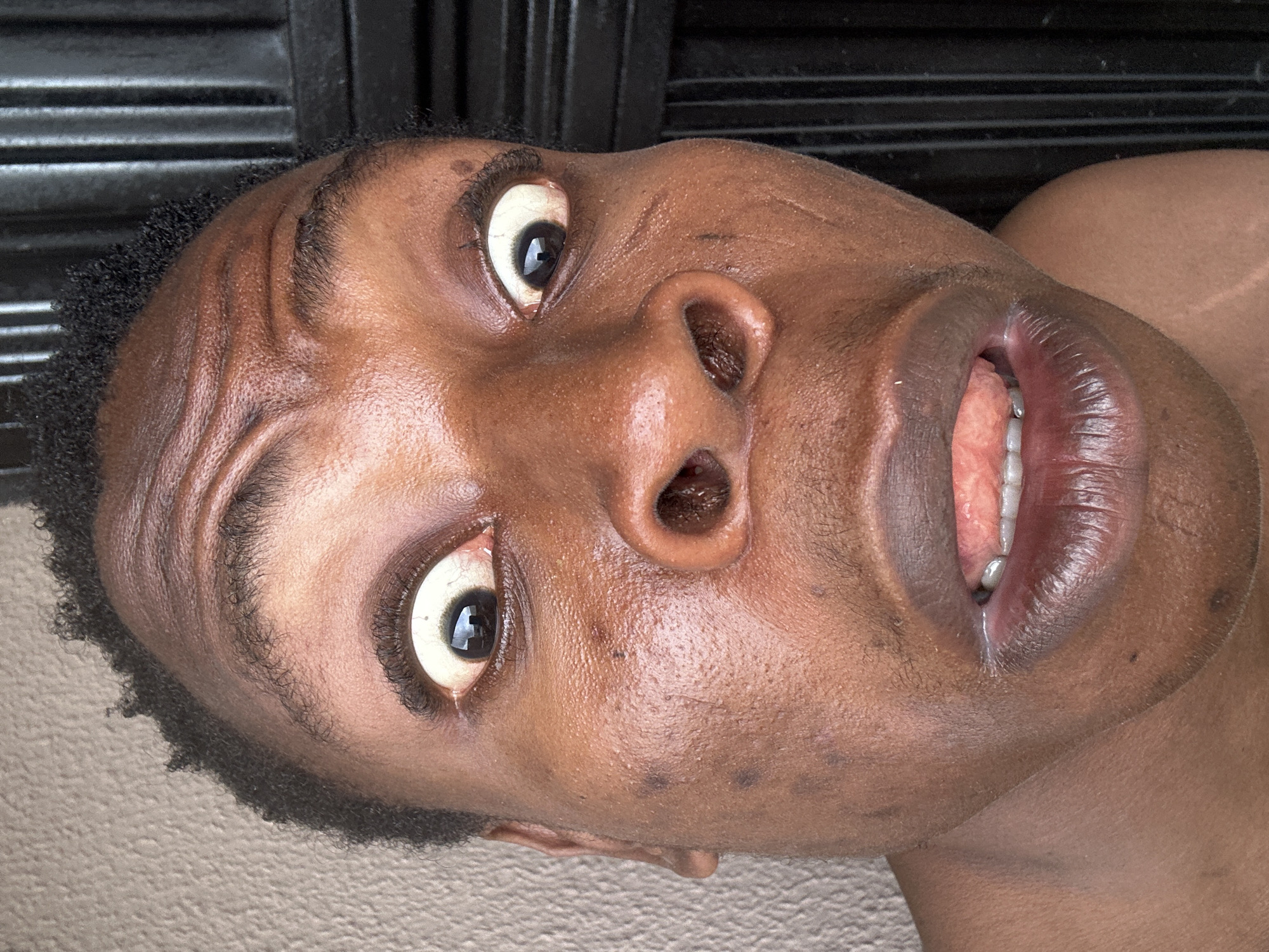 A close-up, tilted shot shows a Black male's face, looking upwards. His eyes are wide, revealing the whites of his eyes. His mouth is slightly open, showing his teeth and tongue. The skin is brown and appears slightly moist. The background is blurred but appears to consist of dark, possibly metallic, horizontal lines above, and a textured surface below. The location is unknown.