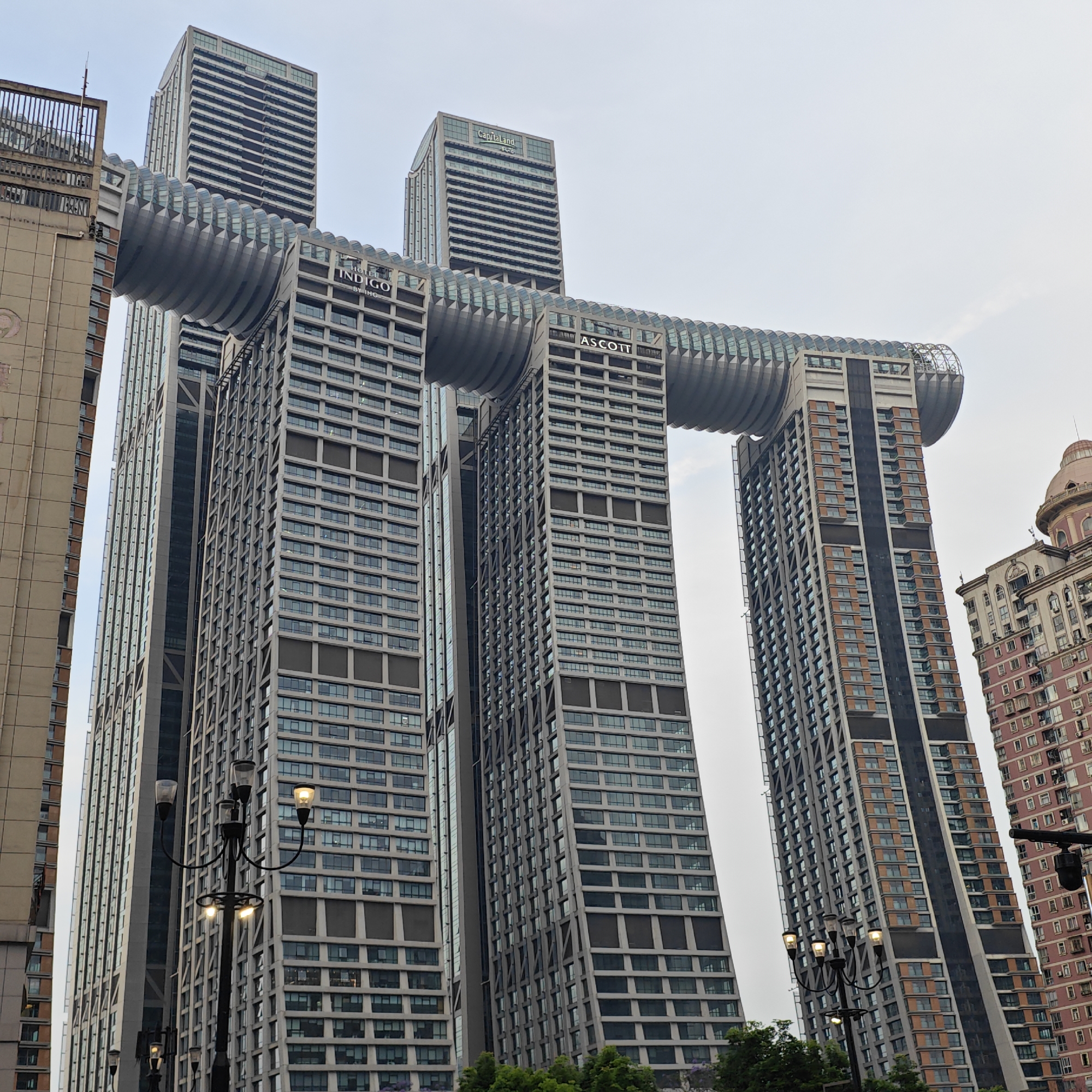 The image depicts the Raffles City Chongqing complex in Yuzhong District, China, under an overcast sky. Three skyscrapers are prominently connected by a large, curved horizontal structure that houses the "Crystal" sky bridge. The buildings are predominantly gray and feature repeated rectangular windows. The names "INDIGO" and "ASCOTT" are visible on the structures. Street lamps with illuminated bulbs stand in the foreground. Older, lower-rise buildings flank the skyscraper complex. The environment appears urban, with some greenery visible near the base of the buildings. There are no people visible.
