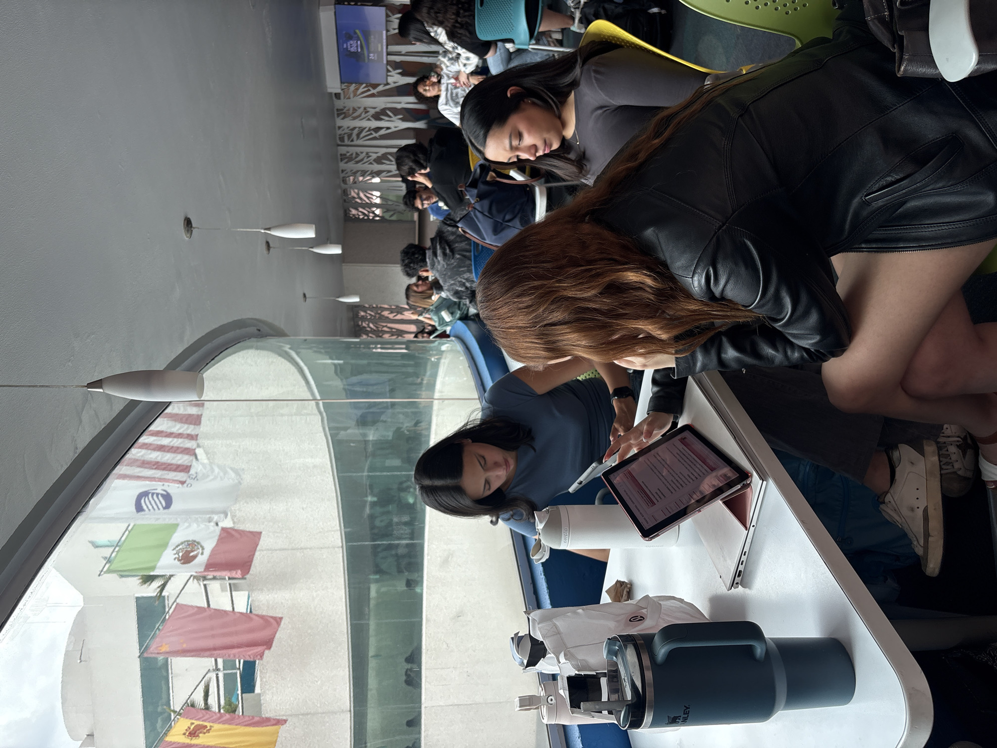 The image shows a group of people sitting at a long, curved table in what appears to be a common area. Several individuals are focused on laptops and tablets, suggesting they are working or studying. Flags of various countries, including the United States, Mexico, and Spain, are visible outside a large window, suggesting an international setting. Based on the flags and the setting, it is possible the location is Universidad de las Américas Puebla (UDLAP) in Heroica Puebla de Zaragoza, Mexico.