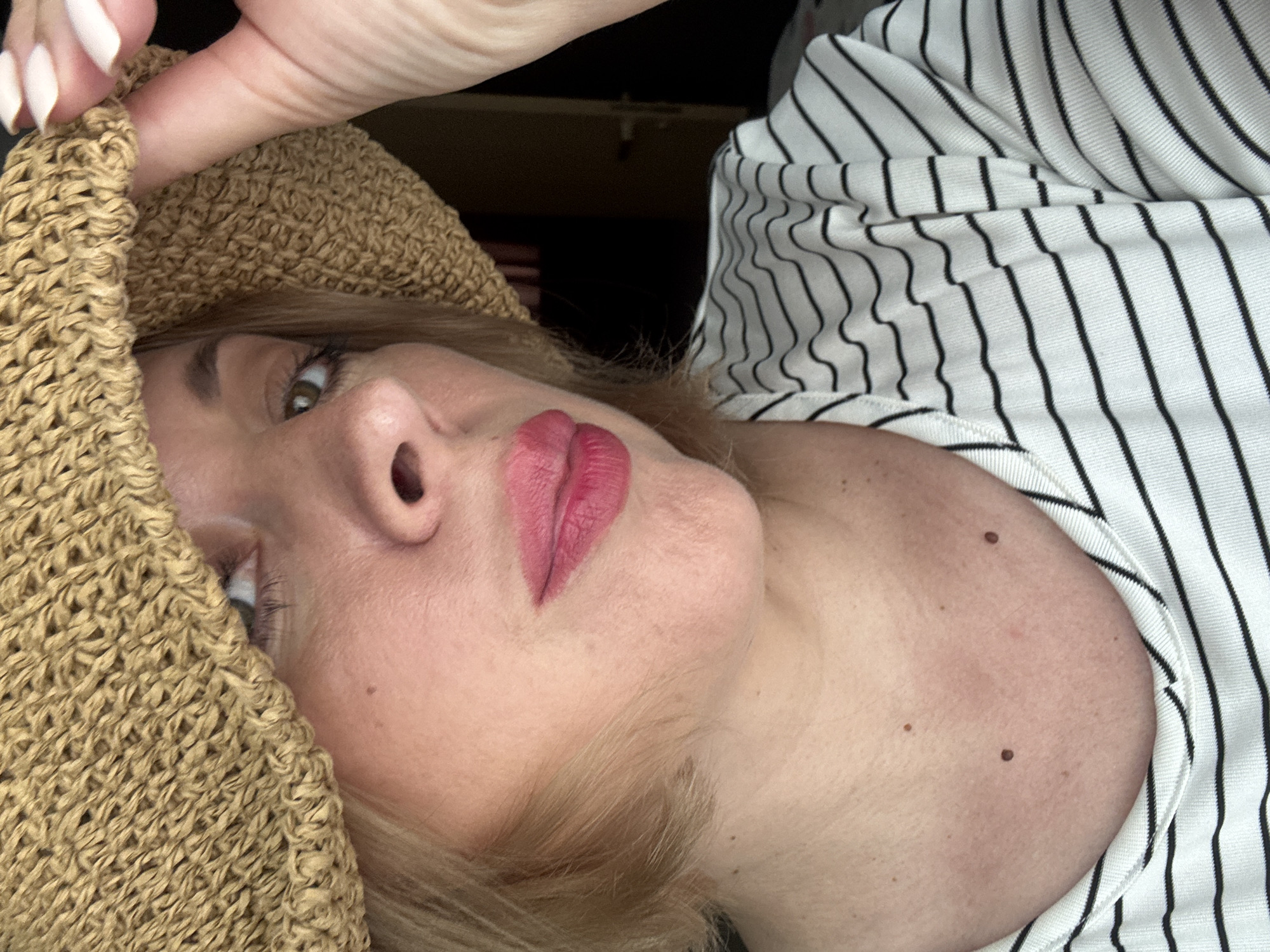 A close-up, eye-level, medium shot captures a fair-skinned person from the chest up, wearing a straw hat and a black and white striped t-shirt. The hat, tilted to cover the top of the head and partially obscuring the face, casts a shadow. The person's left eye is visible, with light brown irises. Their lips, painted with pink lipstick, are slightly parted. The person's right hand, with light-colored fingernails, is raised to the brim of the hat. The background is dark and out of focus. The image does not contain information about the location of Sochi, Russia.