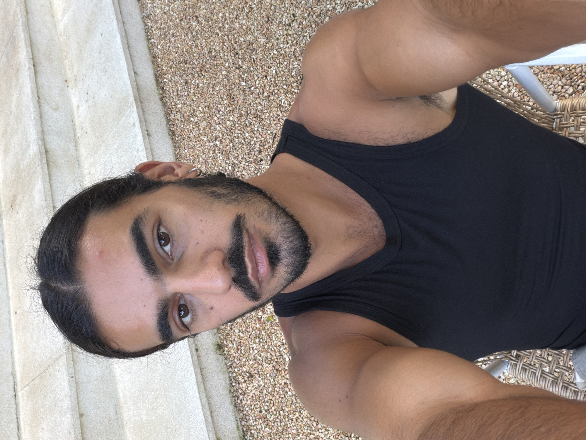 A man with dark hair pulled back in a bun and a mustache and beard is taking a selfie outdoors. He is wearing a black tank top. Behind him, there are light-colored stone steps and a surface covered in small, light-colored pebbles. He is seated on a chair with a woven texture. The image is taken from a high angle, looking down at the man. The setting appears to be a well-maintained outdoor area, possibly a patio or garden.