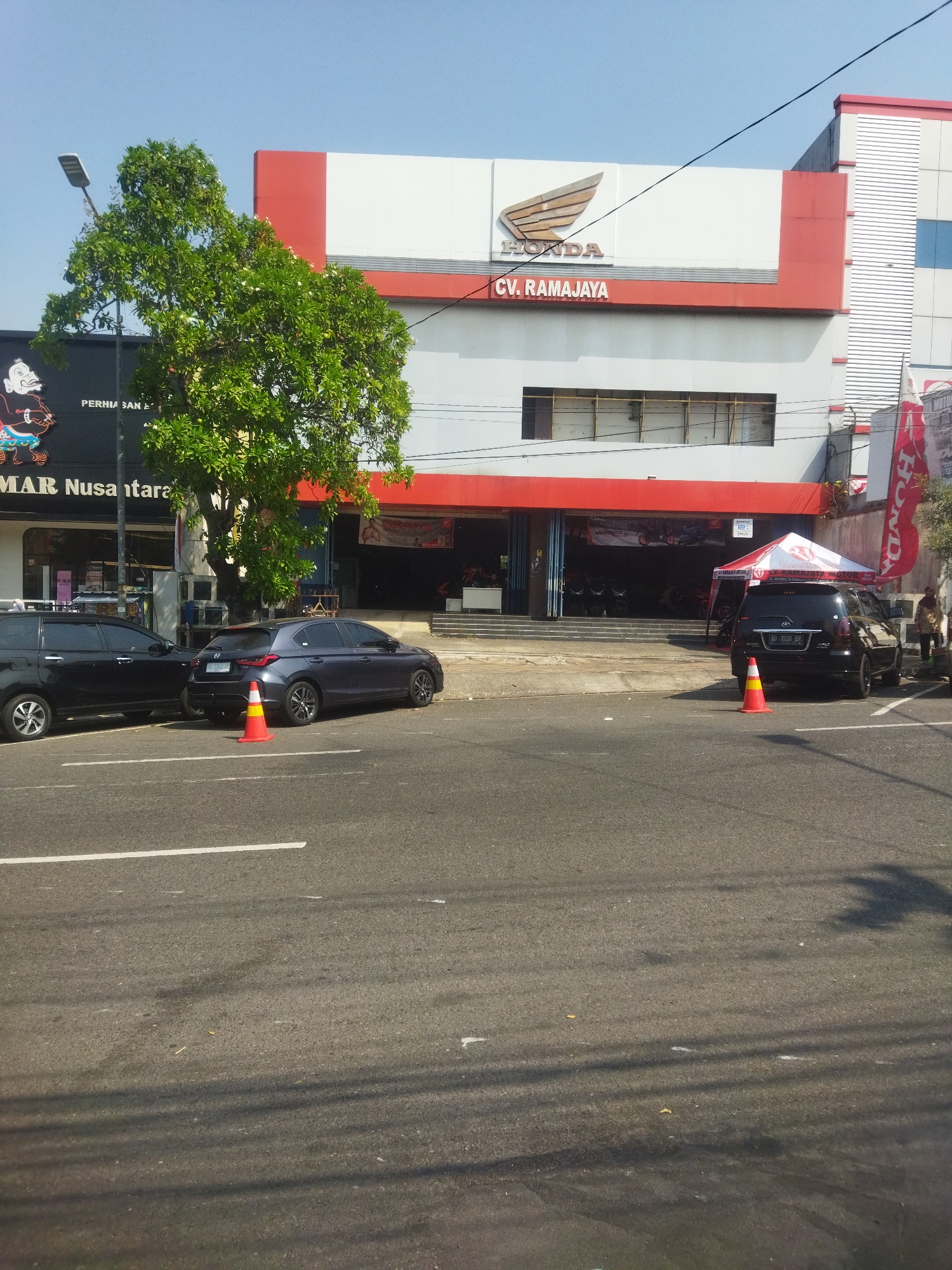 The image shows the exterior of a Honda dealership, identified as CV. RAMAJAYA, located in the city of Wonogiri, Indonesia. The building has a modern facade with red and white accents. In front of the dealership, there are several vehicles parked, including a black SUV and a dark gray hatchback. A red and white traffic cone is placed on the road near the parked vehicles. A large tree with green foliage stands to the left of the building. The sky above is clear and blue. The street in front of the dealership is asphalt with white lane markings.