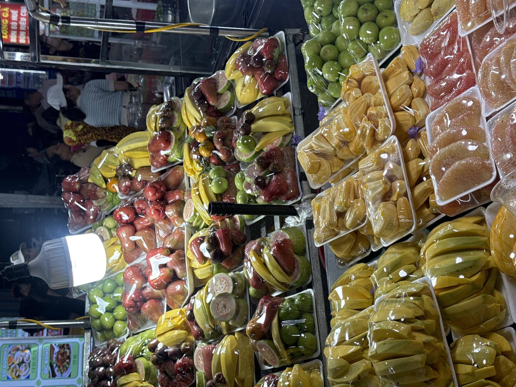 This image shows a fruit stand in Da Lat, Vietnam, filled with neatly packaged fruits like guava, mango, jackfruit, and pomelo, illuminated by an LED bulb. The fruit is arranged on white plastic trays and wrapped in plastic film. In the reflection above the fruit, there is an individual wearing a striped shirt and lying prone. A sign with Vietnamese text "SO DƯƠNG", "ÓC ĐÔNG", and "LONG GÀ" is visible near the bottom left.
