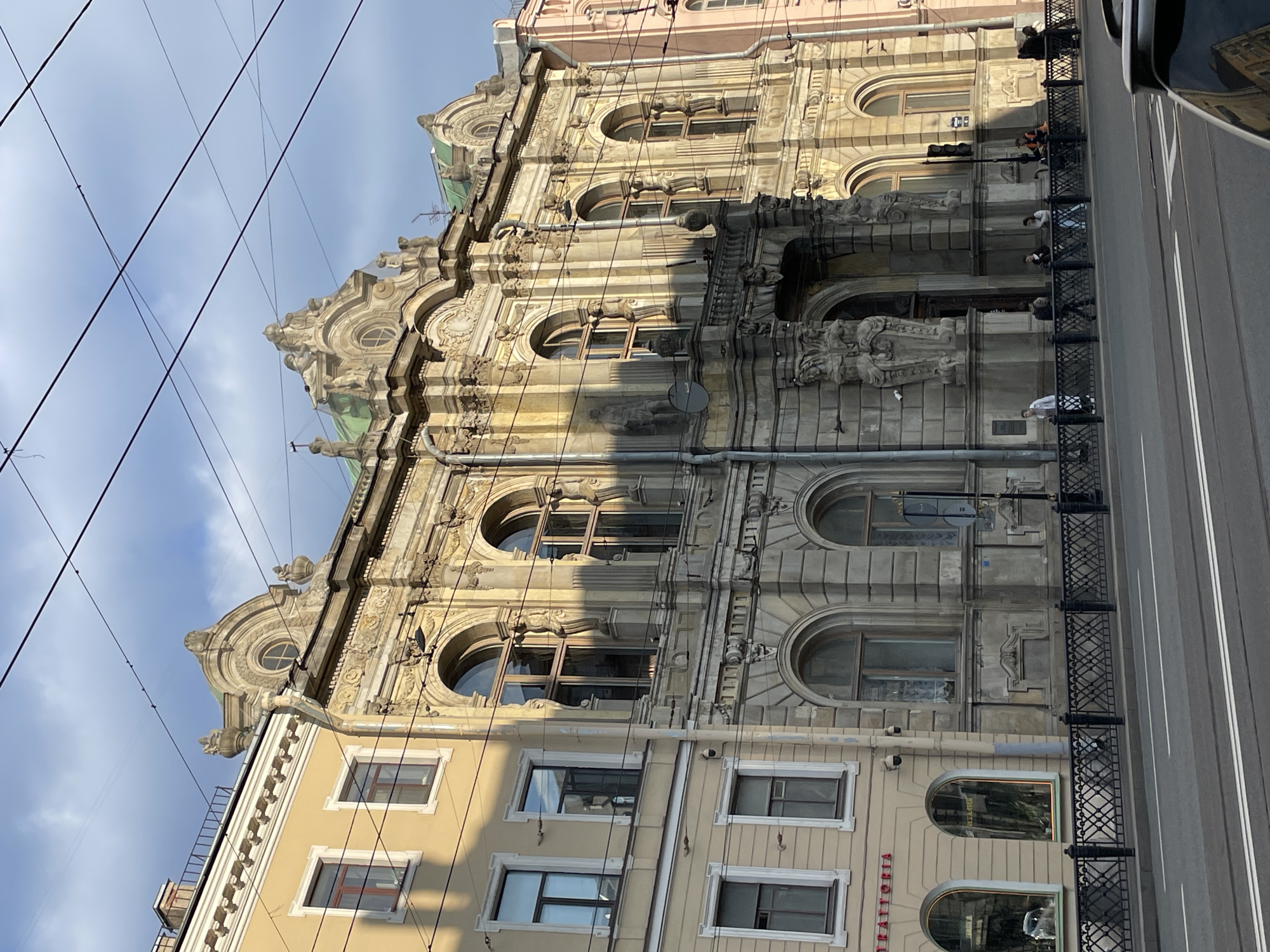 The image depicts the Eliseev Emporium building located on Nevsky Prospekt in Saint Petersburg, Russia. The building features elaborate Art Nouveau architecture with arched windows, ornate facades, and sculptures. A street runs alongside the building with a black metal fence separating the sidewalk from the road. People are visible standing along the sidewalk. Overhead power lines are visible against a backdrop of a partially cloudy sky. The photograph is taken at an angle, looking upwards towards the building.