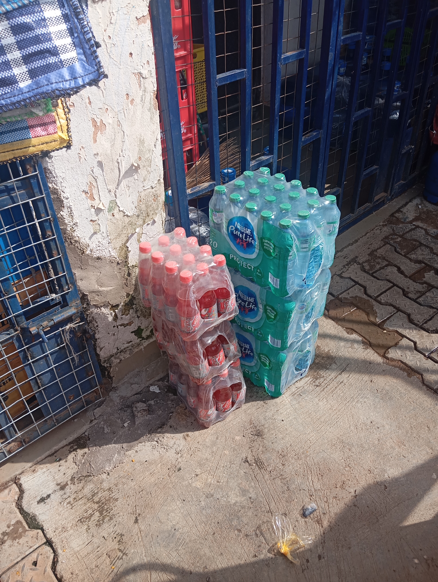 The image depicts an outdoor scene in Gura Top, Nigeria, showcasing stacks of bottled beverages and a weathered building facade. In the foreground, two large stacks of plastic-wrapped bottles are placed on a concrete surface. The stack on the right consists of what appears to be Nestle Pure Life bottled water, with green caps and prominent labeling. The stack on the left contains bottles of red liquid, possibly a soda or juice, with red caps, also shrink-wrapped. Adjacent to the stacks, a blue metal grate structure is visible, possibly part of a stall or shop. The background reveals a textured, distressed wall, painted in a pale, peeling color, with glimpses of red and yellow crates behind a blue metal barred enclosure. A blue and white checkered fabric item is partially visible hanging on the wall. The ground is a patterned concrete pavement with some damp patches and scattered debris, including a small plastic bag and an empty plastic bottle. The lighting suggests a sunny day, casting shadows on the ground. There are no people visible in the image.