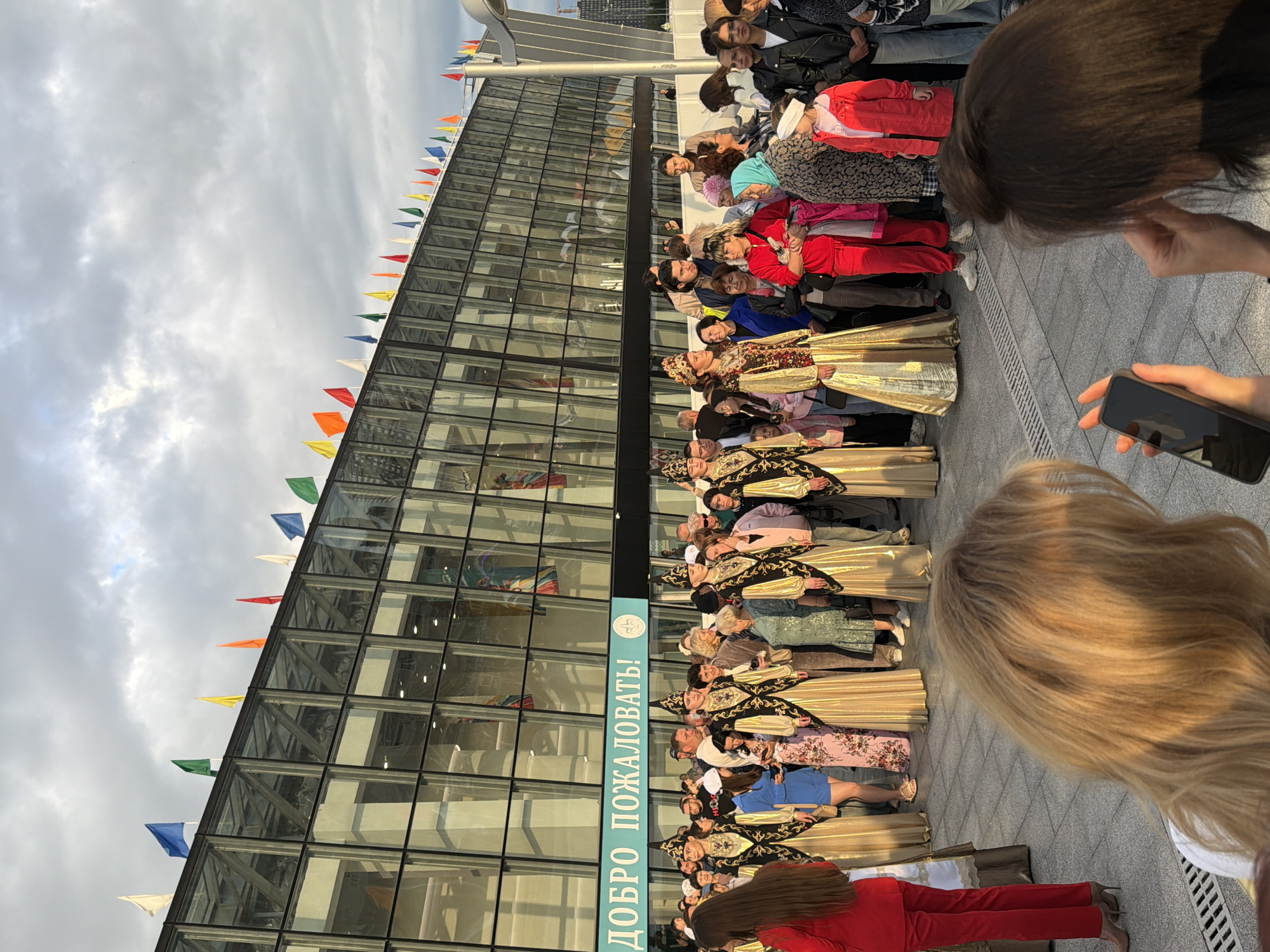A group of people stand in front of a modern building with a glass facade, adorned with numerous colorful flags along its roofline. Several individuals in the foreground are wearing traditional gold and black embroidered outfits with conical hats, possibly participating in a cultural event or performance. To the right, a woman in a red jacket holds a smartphone and appears to be taking a photograph or video. The building features a large sign with text in Cyrillic, which appears to say "ДОБРО ПОЖАЛОВАТЬ!" meaning "Welcome!". The ground is paved with grey tiles. The sky is partly cloudy with a warm, golden light suggesting it might be late afternoon or early evening.