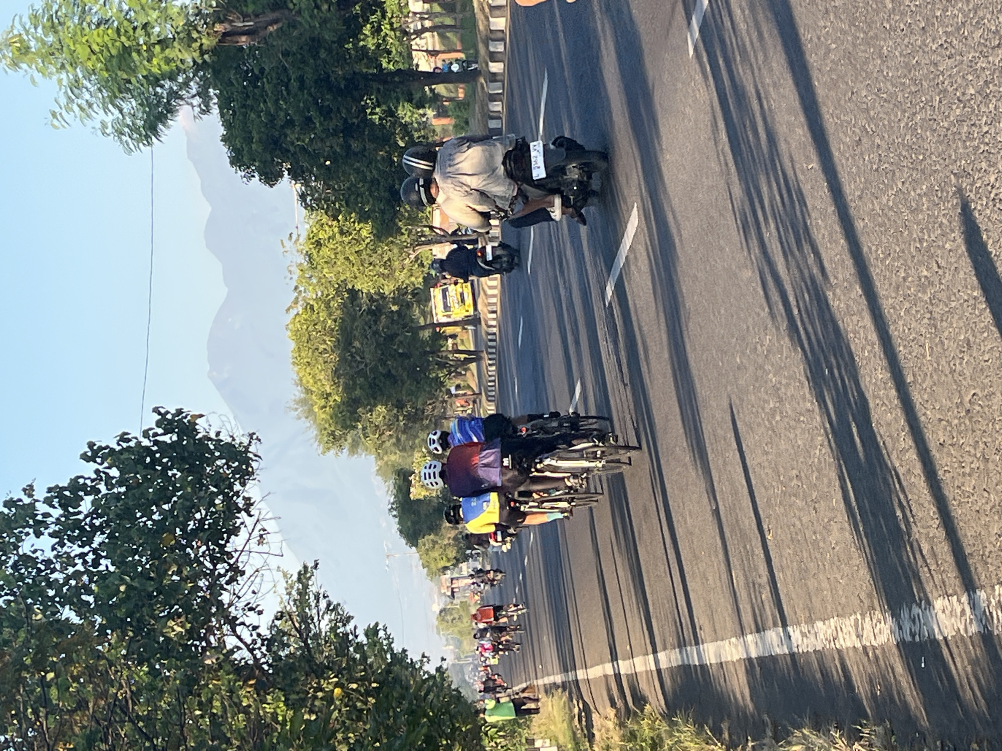 The image depicts a street scene in Siring, Indonesia, with a clear blue sky overhead and distant mountains. The sun is casting long shadows across the asphalt road, indicating it is either early morning or late afternoon. A group of cyclists are riding in formation on the left side of the road, many wearing helmets and brightly colored cycling jerseys. On the right side of the road, a motorcycle carrying two people, both wearing helmets, is also in motion. Several other cyclists and a yellow vehicle are visible further down the road. The road is lined with trees and vegetation on both sides, and a sidewalk with a concrete barrier is present on the right. The overall impression is one of a group activity or commute taking place on a sunny day in a semi-urban or suburban area.