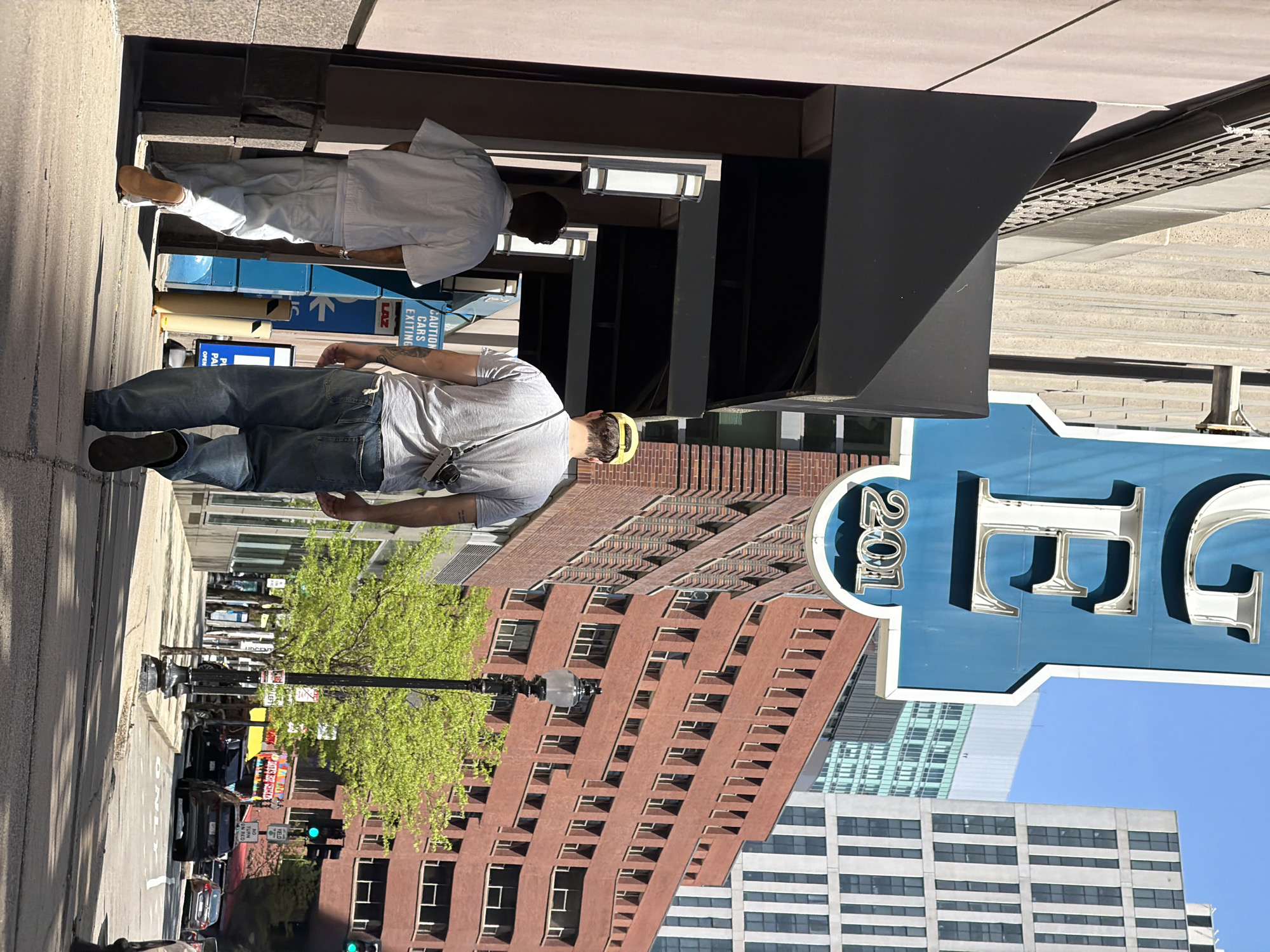 This image shows a street scene in Boston, Massachusetts. Two men are walking on the sidewalk in front of a building. The first man is wearing a light grey t-shirt, jeans, and brown shoes and has a yellow headband. The second man is wearing white pants and a white dress shirt. The street contains vehicles and a tree. The large sign on the right says "201 E". Buildings of varying heights and architectural styles fill the background. The sky is blue.