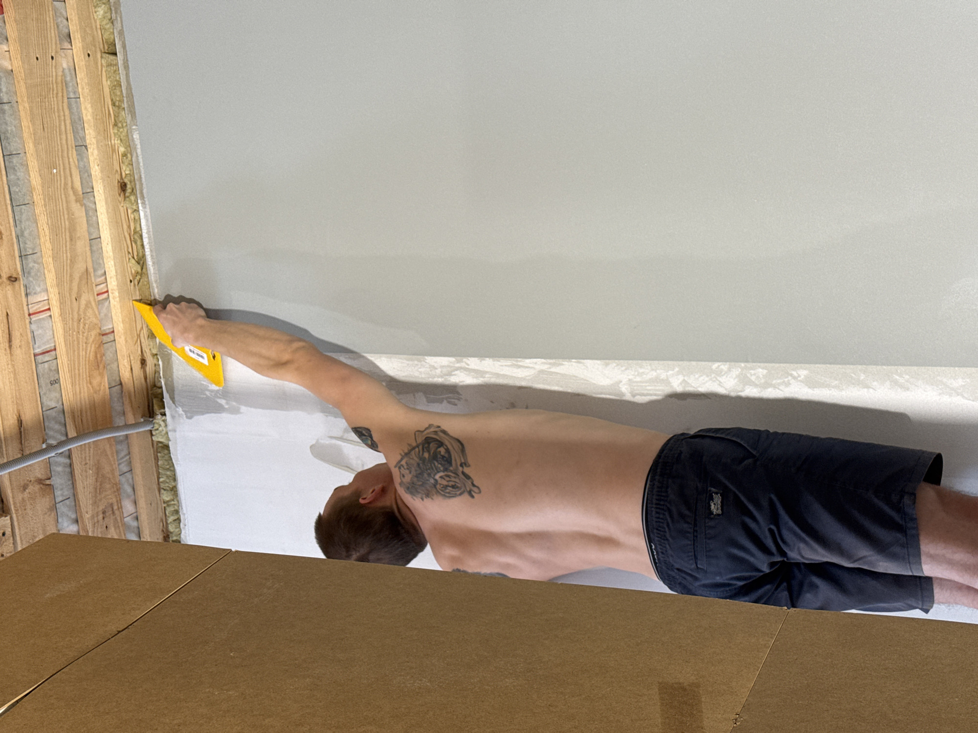 A shirtless man wearing dark shorts is applying spackle to a wall using a yellow trowel. The wall appears to be newly constructed with wooden framing and insulation visible along the left edge. The man has tattoos on his back and shoulder. The scene includes a gray wall, white spackle, and brown cardboard on the foreground. The photograph was taken at Chaikovsky, Russia.
