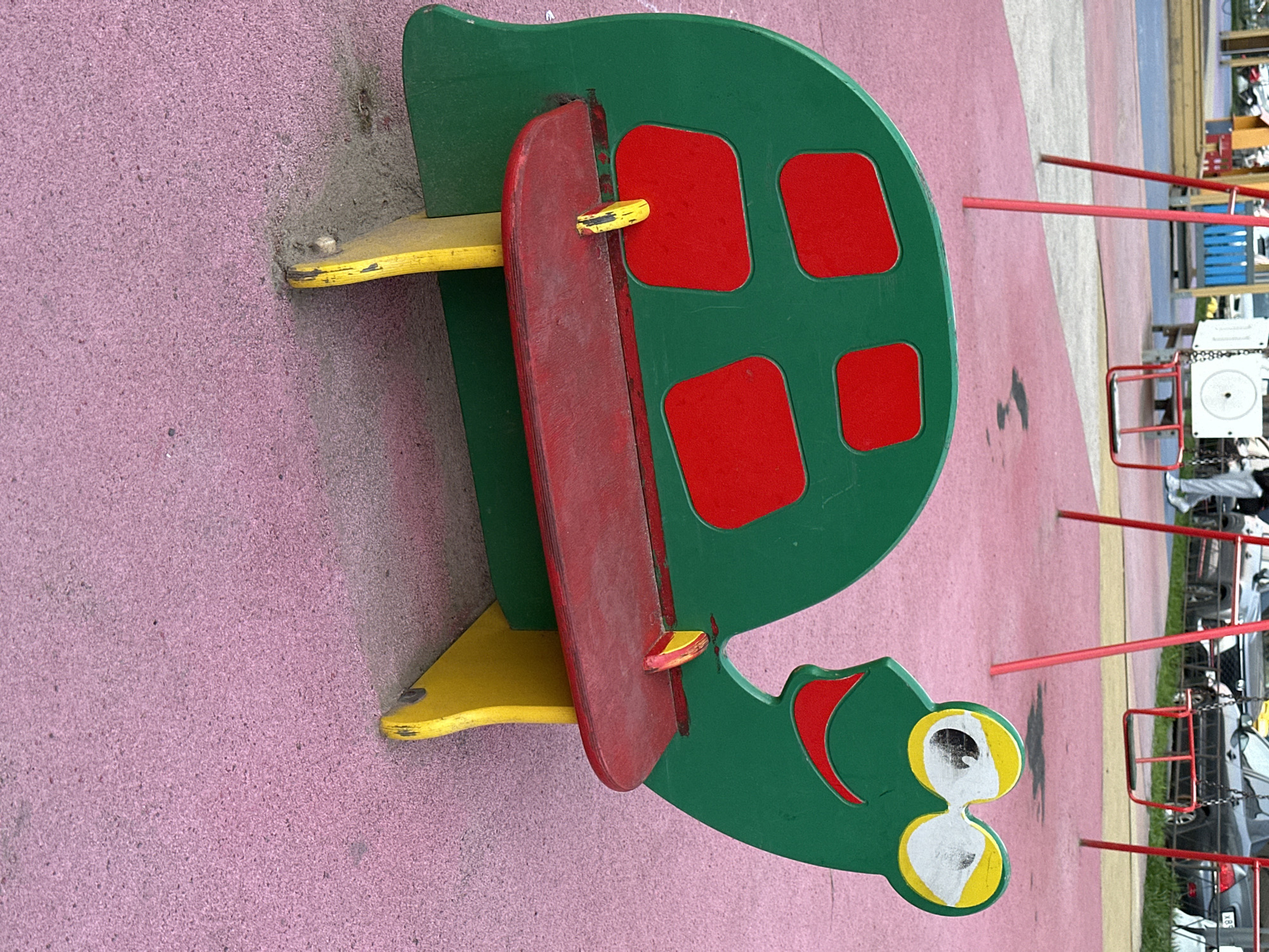The image shows a playground in Kudrovo, Russia. The main subject is a green and red turtle-shaped spring rider toy on a pink surface. The toy has a green shell with four red squares, a red seat, and a green head with yellow and white eyes. The base and supports are yellow. In the background, a playground structure with red bars and other equipment can be seen. The environment appears to be an outdoor play area within a city park or recreational space. Cars are visible in the far background.