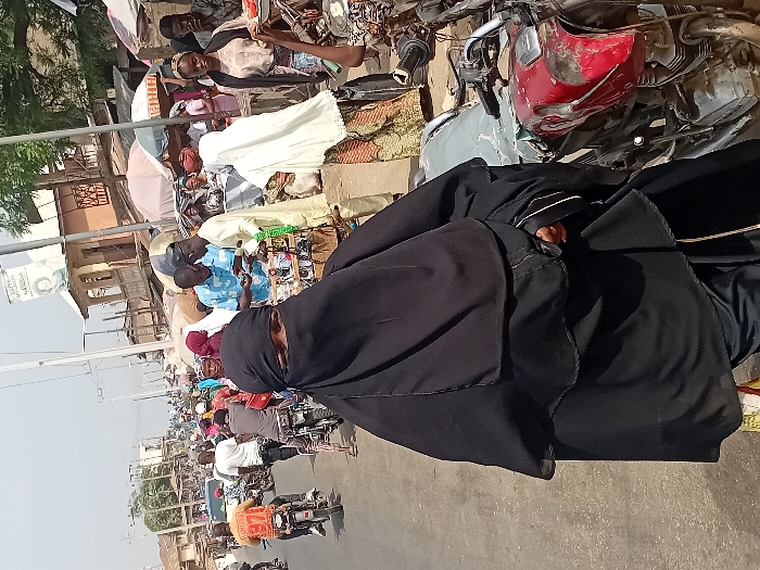 This image depicts a bustling street scene in Ogaminan, Nigeria, during the daytime under a clear sky.
In the foreground, a person is prominently featured, wearing a black niqab that covers their entire face except for their eyes, and a matching black abaya. They appear to be seated on a motorcycle, with a red motorcycle fender or fuel tank partially visible behind them. Their right hand, partially covered by the abaya, is holding onto something, and a wrist accessory is visible.
The background reveals a vibrant and crowded environment. To the left of the main subject, a busy market area is visible, teeming with people dressed in various attire, including traditional garments. Vendors are present, some behind stalls or displaying goods on the ground, with a light-colored umbrella offering shade. Further back, buildings with unpainted brick or concrete facades line the street. A white sign on a pole is discernible, though blurry, with some visible text fragments such as "AD", "OPO", and "N4,000".
To the right of the main subject and extending into the mid-ground, a busy street is packed with numerous individuals, many on motorcycles, suggesting heavy traffic and constant movement. The ground is paved, indicative of a developed road. The overall scene conveys a sense of everyday life and commerce in a lively West African urban setting.