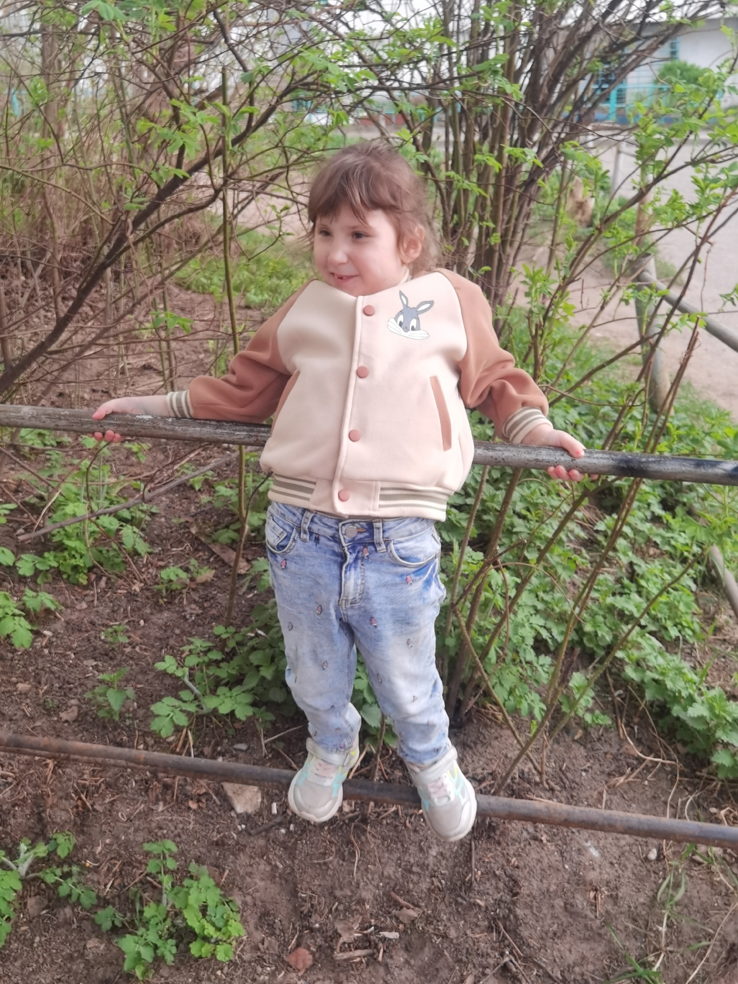 A young girl is standing on a metal railing in what appears to be a park or garden. The girl has light brown curly hair and is wearing a light-colored jacket with brown sleeves and a cartoon rabbit on the front, blue jeans, and sneakers. She is holding onto the railing with both hands, balancing herself. Behind her are bushes with green leaves, and the ground is covered in dirt and small green plants. The location is identified as the city of Chaikovsky, Russia.