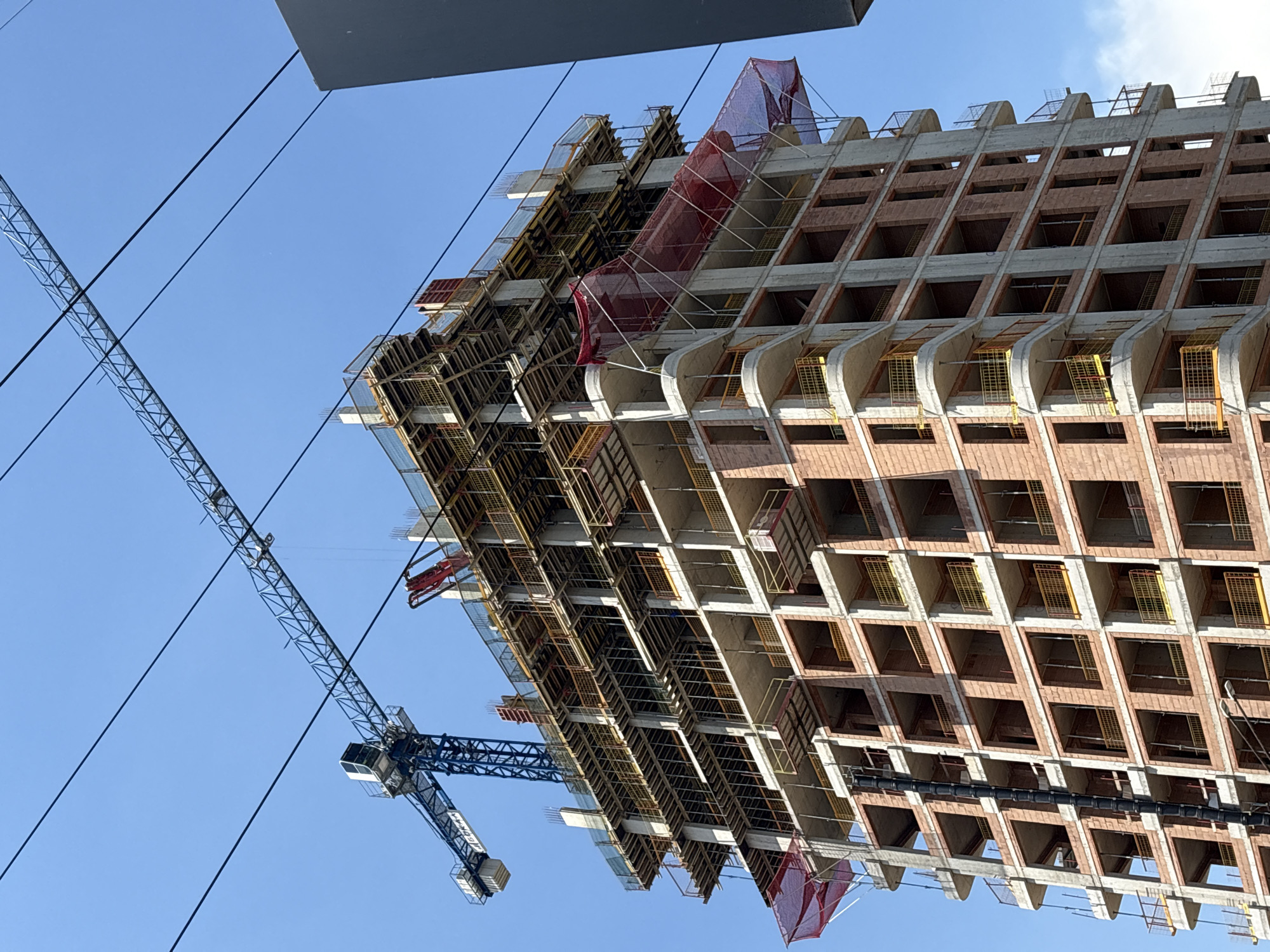 The image shows a tall, unfinished building under construction in the city of Sofia, Bulgaria. A large blue tower crane is positioned to the left of the building, with its boom extending over the top floors. Several power lines are visible in the sky, crossing above the crane and the building. The building itself is made of concrete and brick, with open window frames and scaffolding on some of the upper levels. Red safety netting is draped around parts of the structure. The sky is clear and blue. No people are visible in the frame.