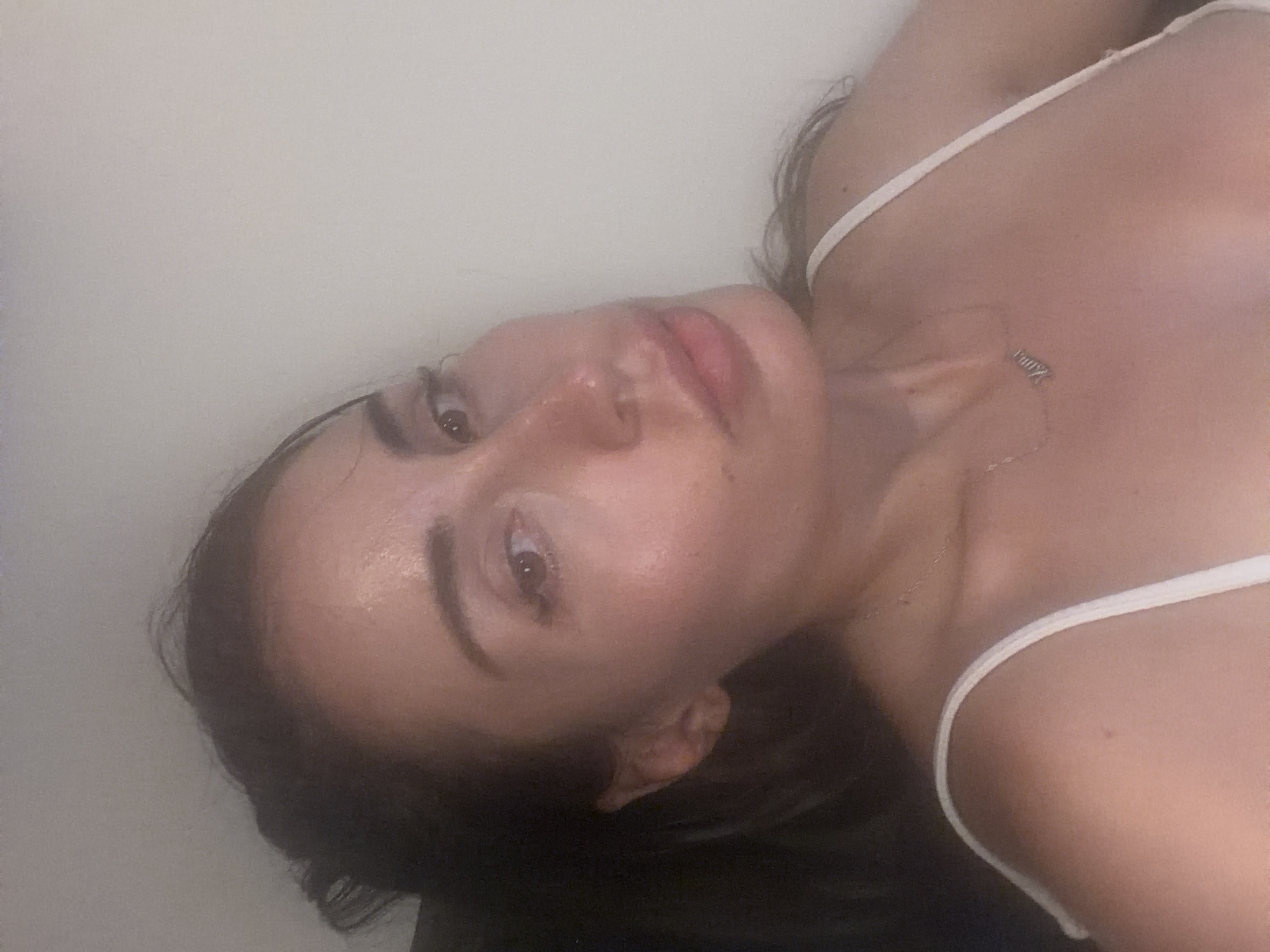 A woman is pictured in a close-up, head-and-shoulders shot. She has dark hair pulled back and is wearing a white camisole with thin straps. A delicate silver necklace with a nameplate hangs around her neck. The background is a plain, neutral wall. The lighting suggests an indoor setting, possibly at night. The image does not contain any identifiable landmarks or features that would suggest a specific location within Tersefanou, Cyprus.