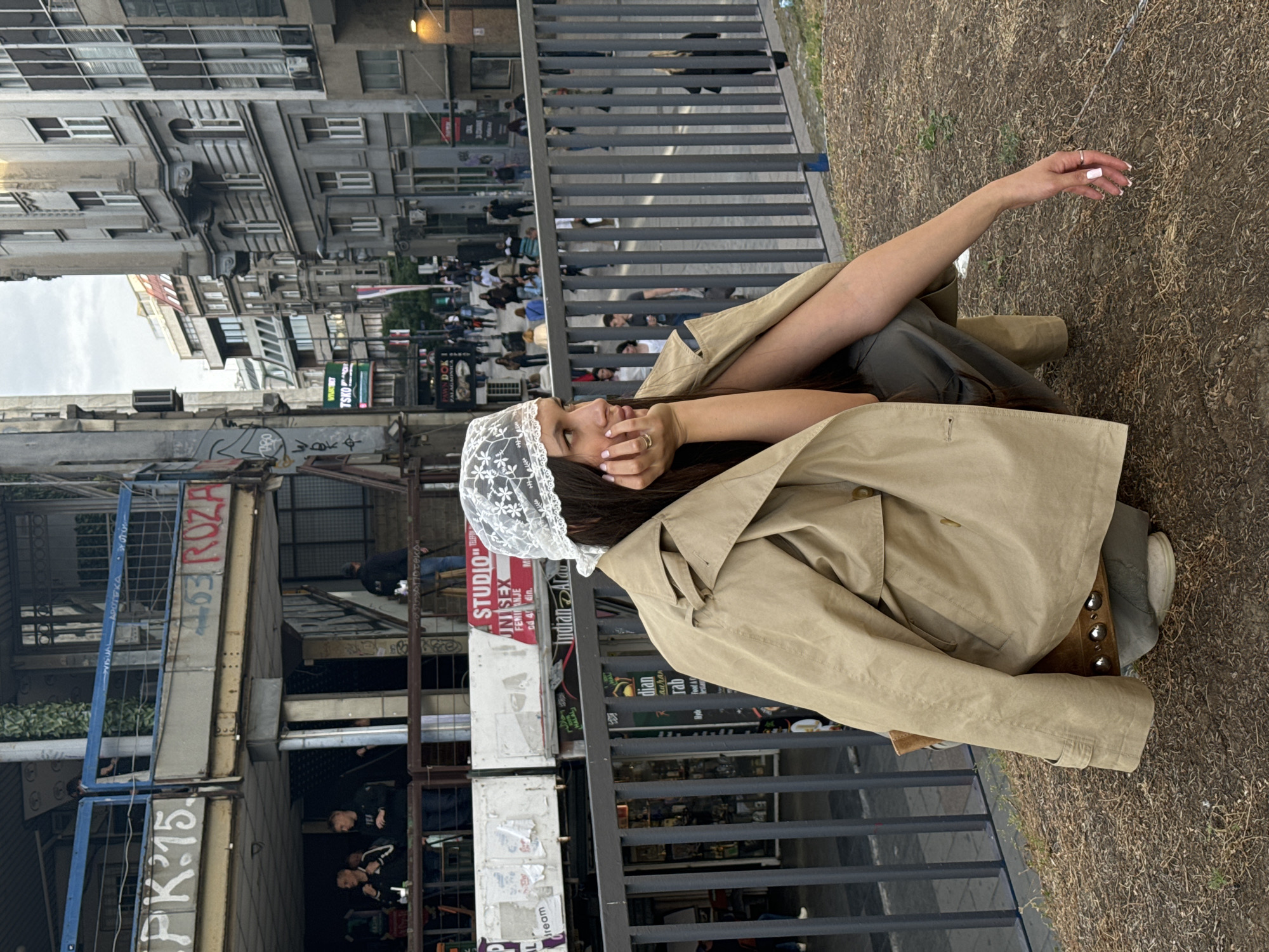 A young woman wearing a tan trench coat and a white lace head covering is reclining on the ground next to a grey metal railing. Her right arm is extended upward. Buildings, sidewalks, and pedestrians are visible in the background. Signage with text in Serbian indicates that the location is in Belgrade, Serbia. Street-level shops and graffiti are visible.