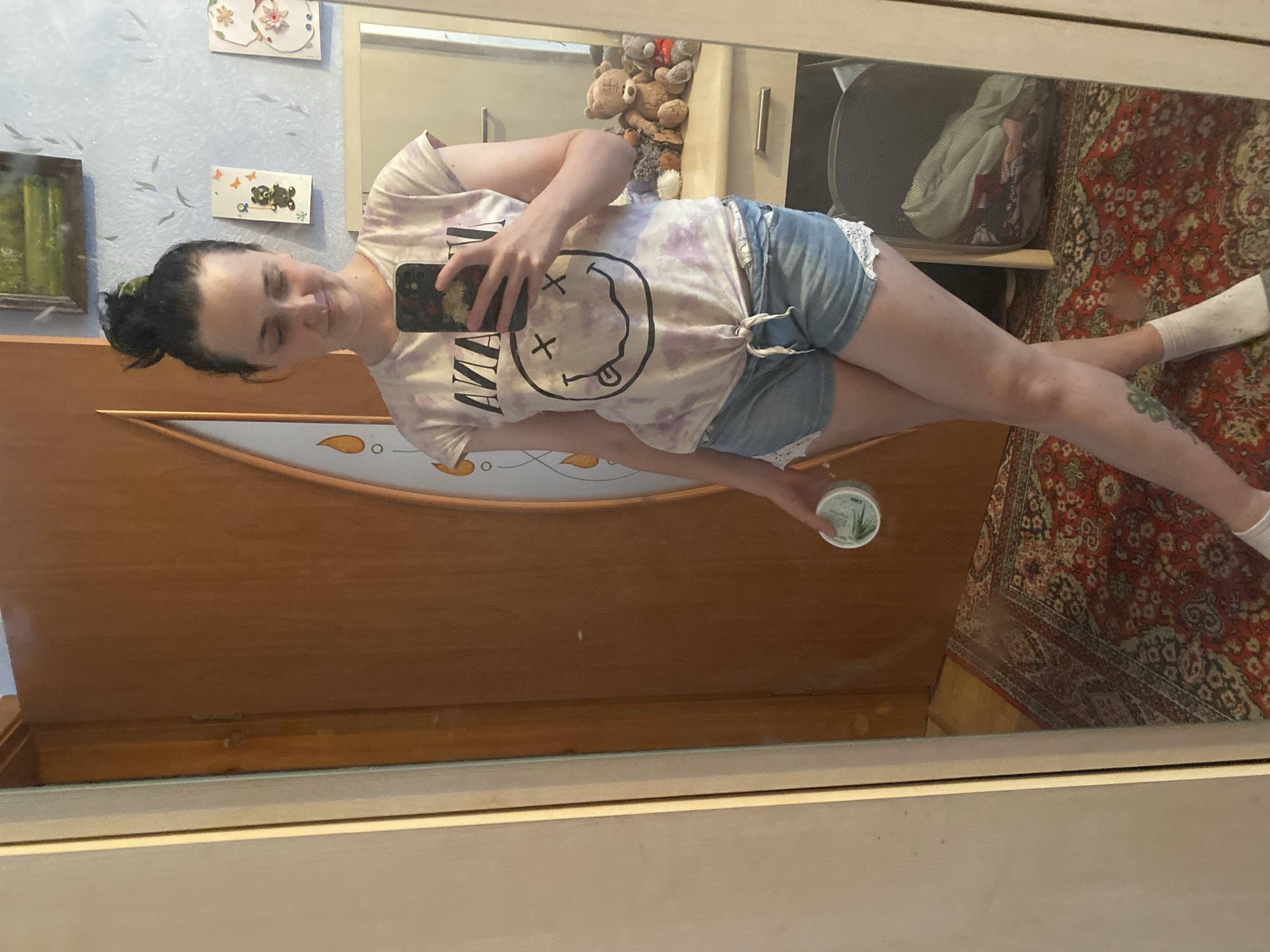 A young woman with dark hair in a bun stands in front of a mirror, taking a selfie with her phone. She is wearing a white t-shirt with a tie-dye pattern and a Nirvana smiley face logo, and denim shorts with lace trim. She is holding a small round container in her right hand. The background shows a wooden door, a patterned rug, a dresser with stuffed animals, and decorative items on the wall. The scene appears to be indoors, possibly in a bedroom or dressing area.